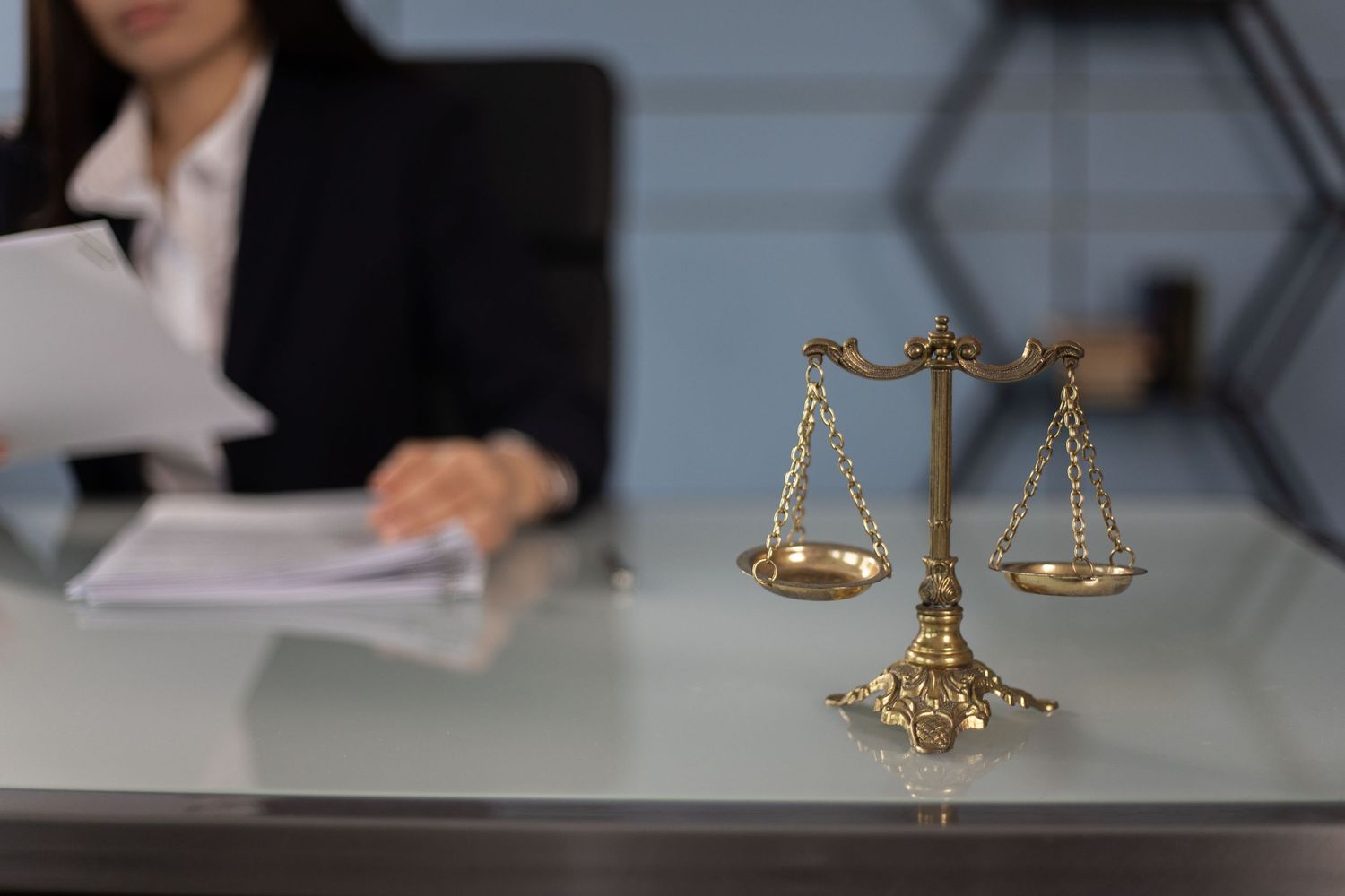Gold scales of justice on glass desk with female lawyer reviewing legal documents.