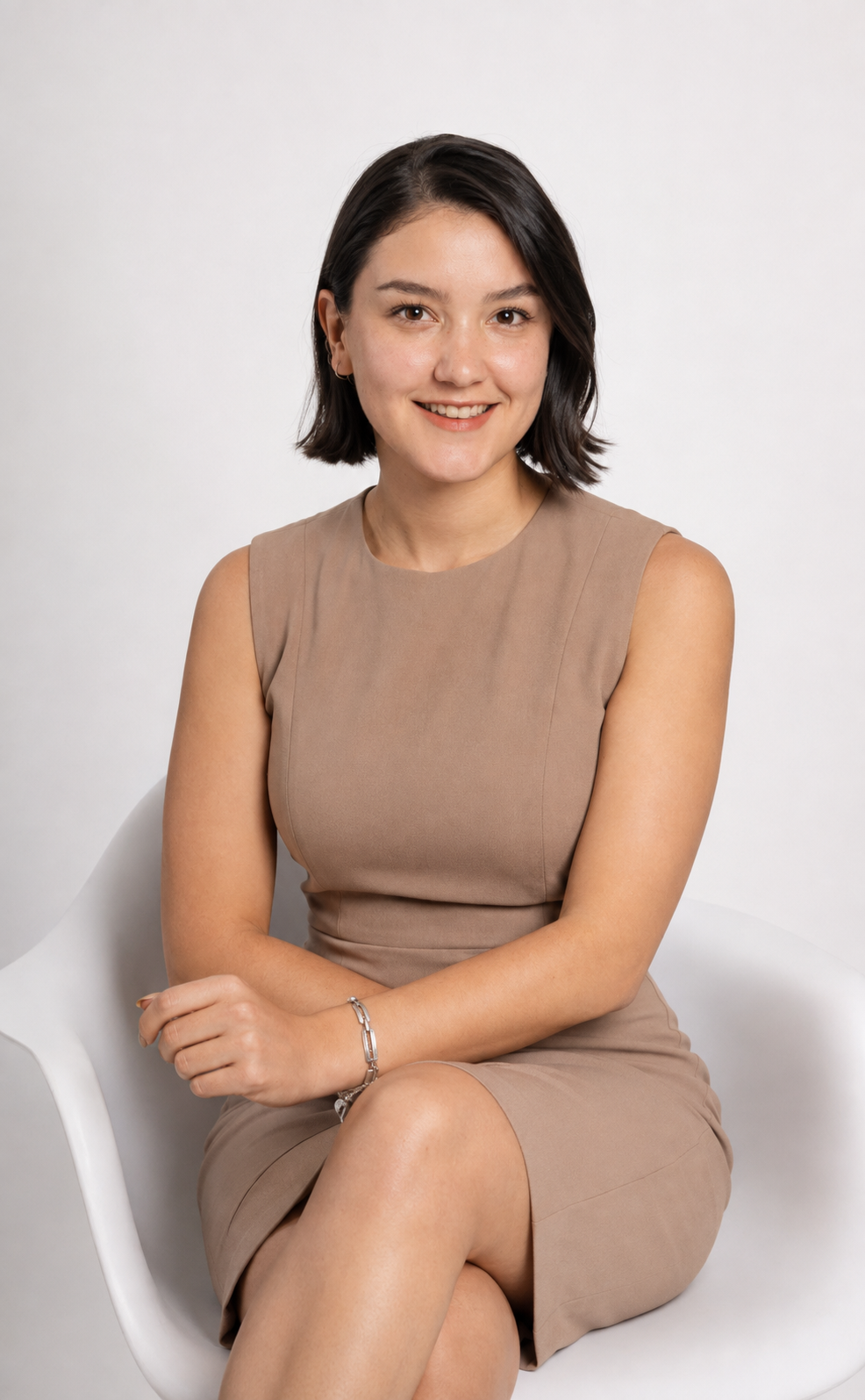Young professional woman in beige sleeveless dress sitting confidently in white chair, smiling warmly against light background.