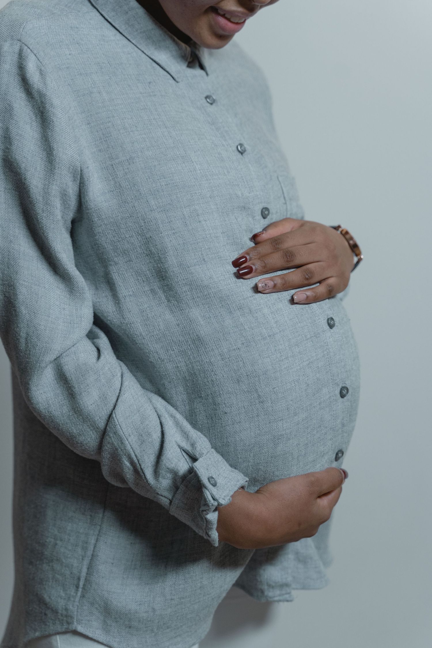 Pregnant woman in a gray linen shirt gently cradling her baby bump against a light background.