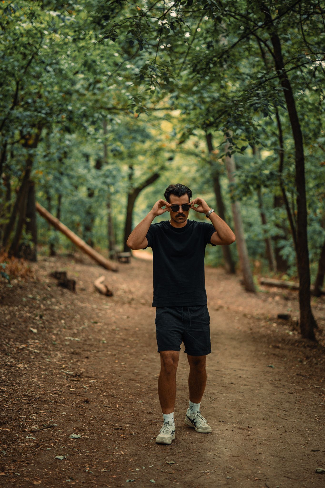 Man in black t-shirt and shorts adjusting sunglasses on a forest trail surrounded by lush green trees.