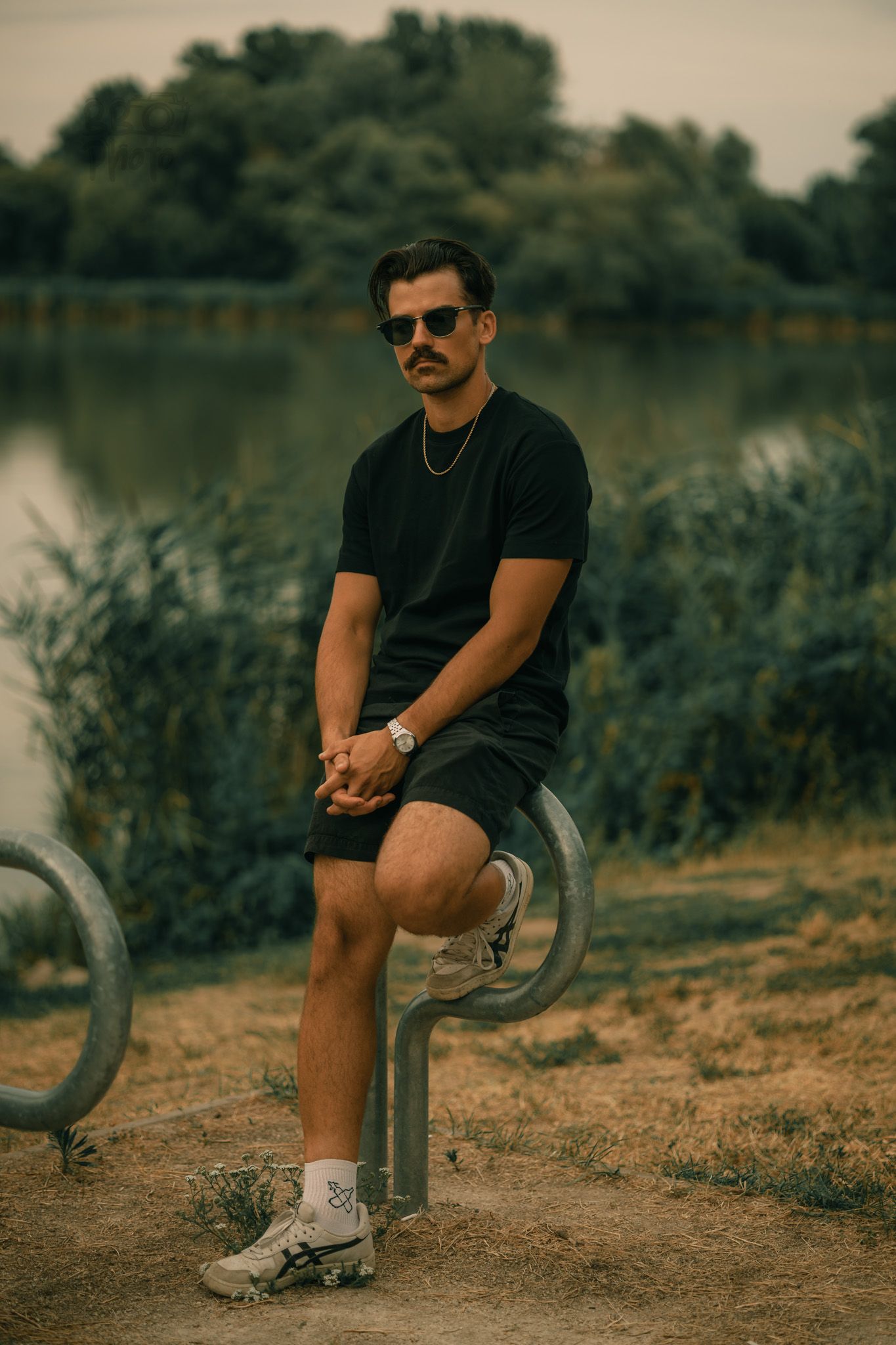 Man in black outfit and sunglasses sitting on metal equipment near a lake with green trees in background.