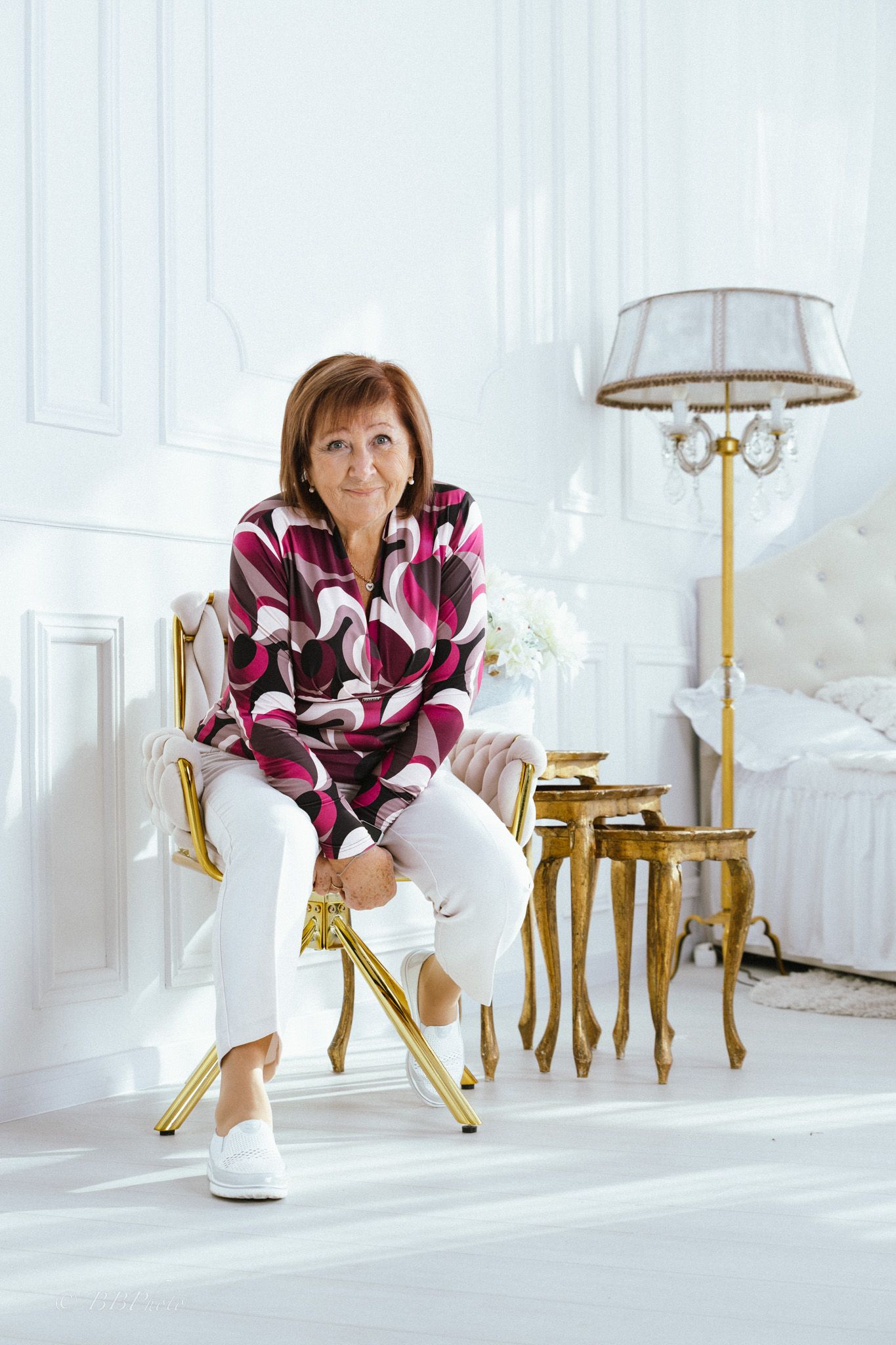 Smiling woman in a burgundy patterned blouse and white pants sits on a gold chair in a bright white room with a floor lamp and bed.