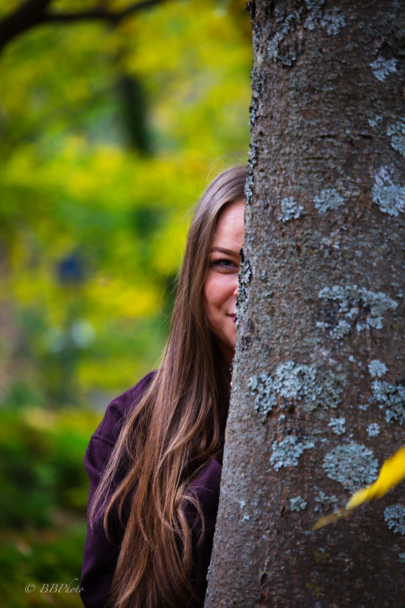 Smiling woman with long brown hair peeks from behind a lichen-covered tree trunk in an autumn forest with blurred yellow and green foliage.