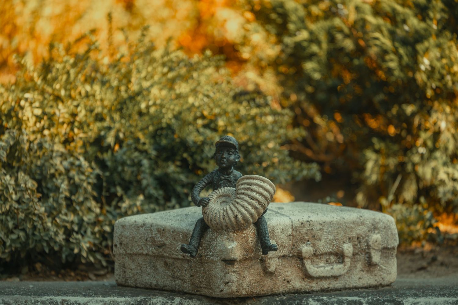 Bronze sculpture of a small figure resting on a stone suitcase surrounded by autumn foliage outdoors.