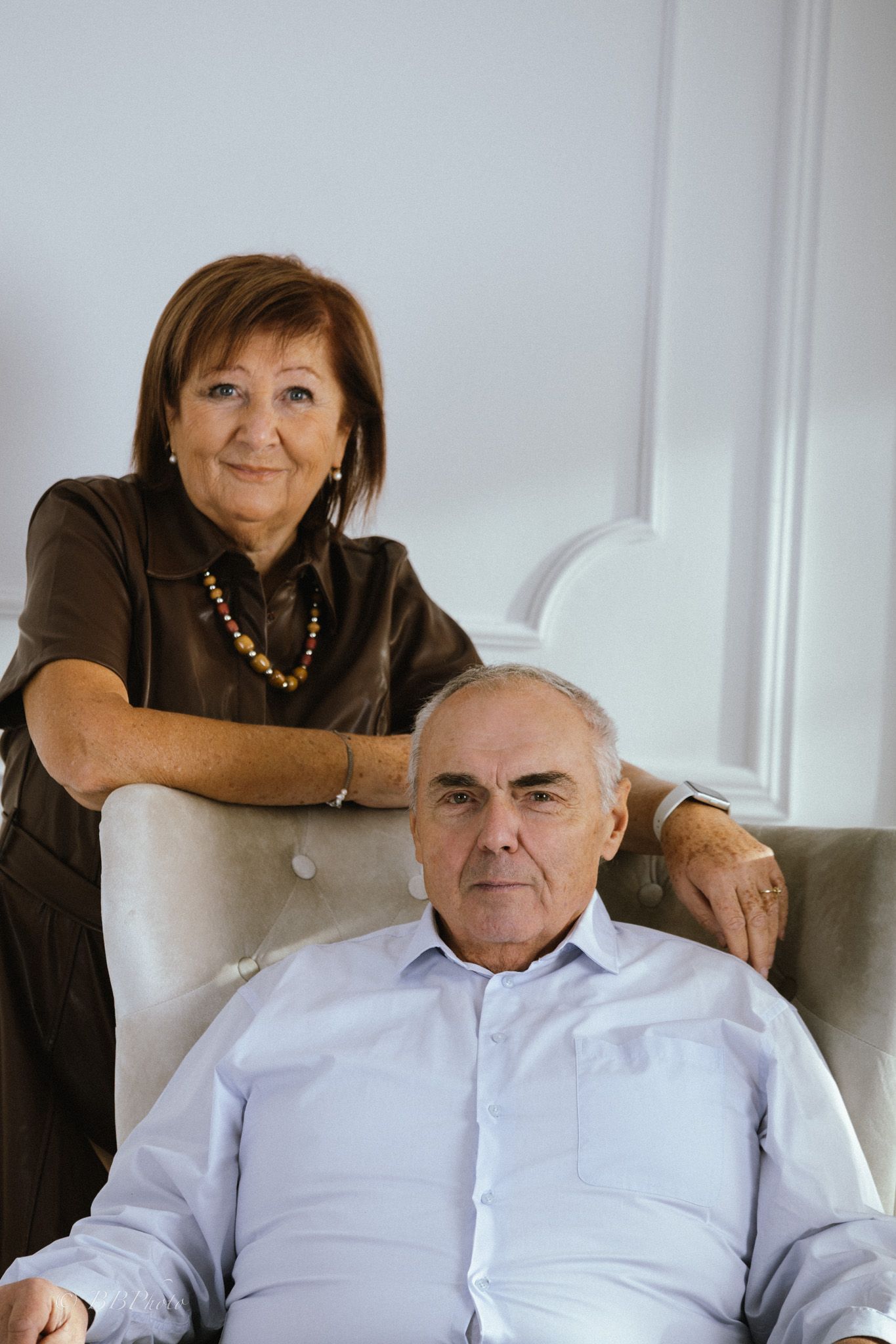 Older woman in a brown blouse and beaded necklace stands behind a seated gray-haired man in a light blue shirt in an elegant armchair.