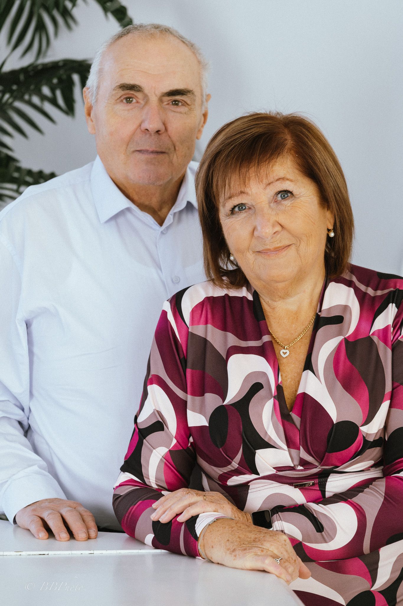 Smiling older couple posing at a white table, the man in a light blue shirt and the woman in a patterned magenta blouse.