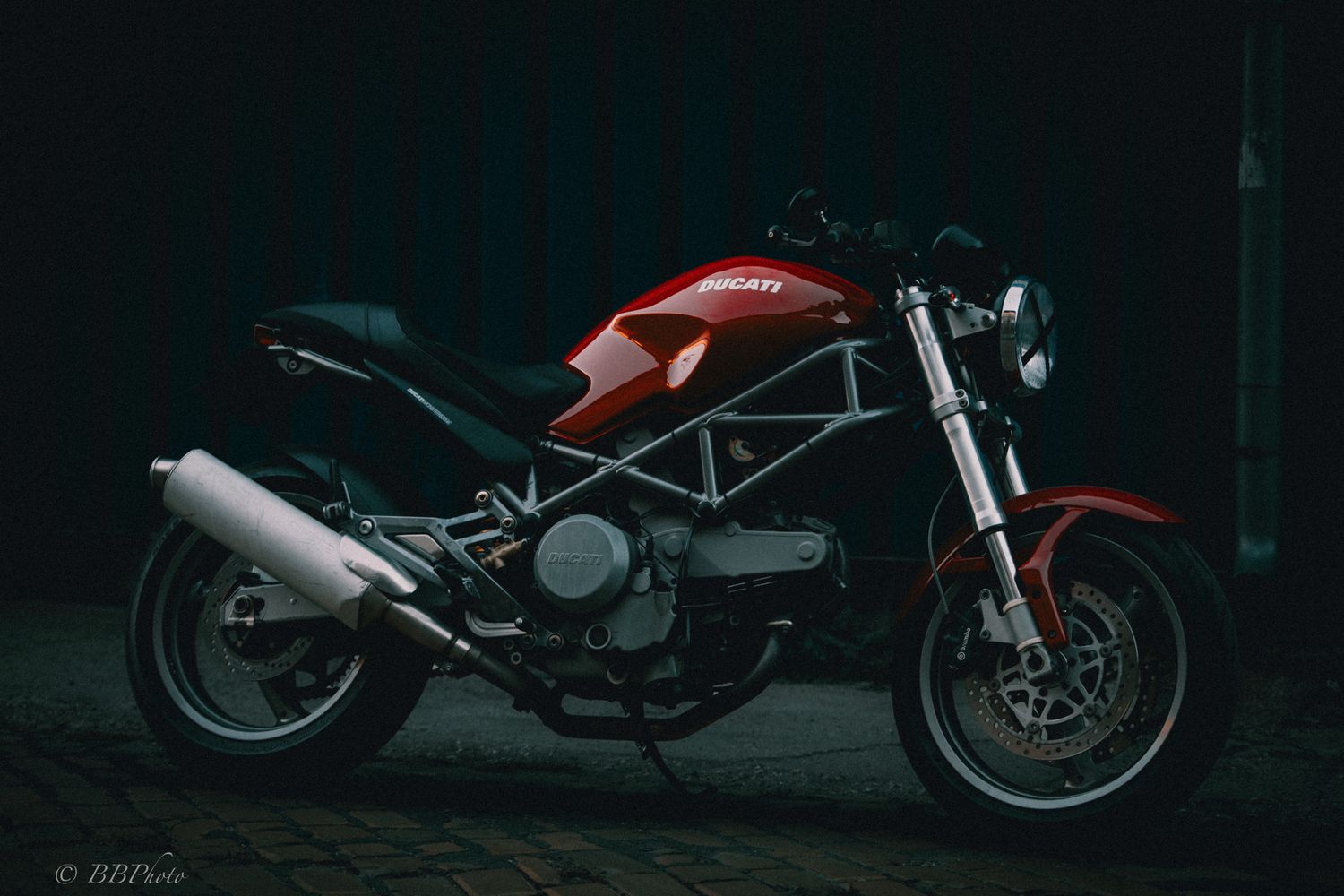 Red Ducati Monster motorcycle with exposed steel trellis frame and chrome exhaust parked on cobblestones against a dark background.