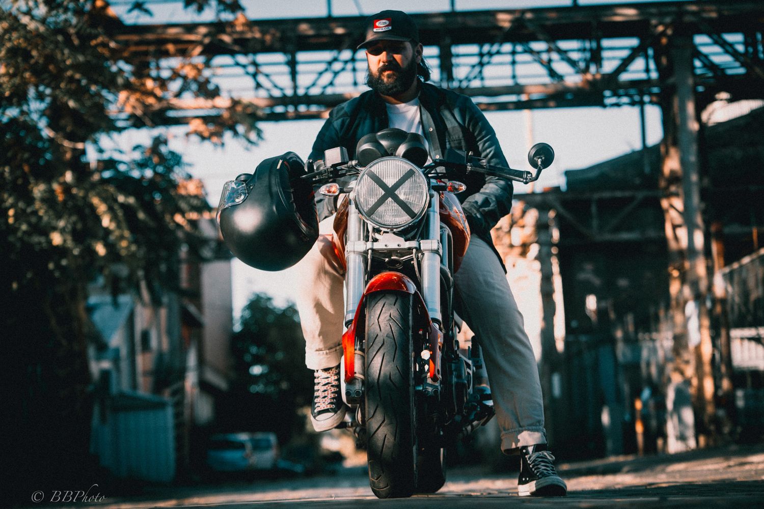 Bearded motorcyclist in a leather jacket and cap sits on a custom red and white motorcycle holding a helmet on an industrial city street.