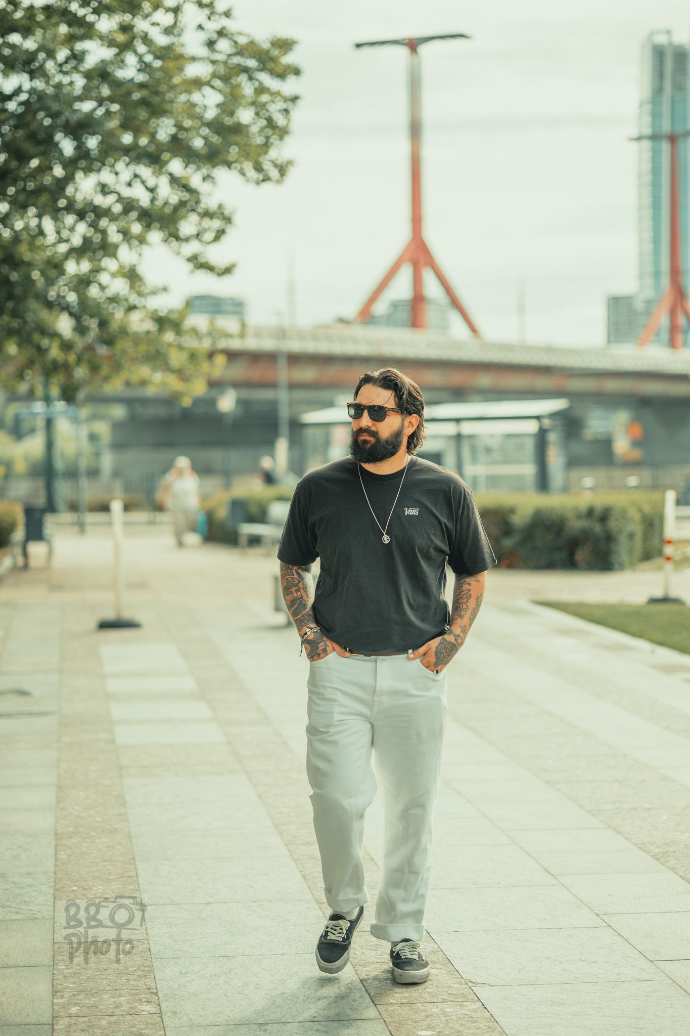 Tattooed man with beard and sunglasses walking outdoors in black tee and gray pants near urban bridge.