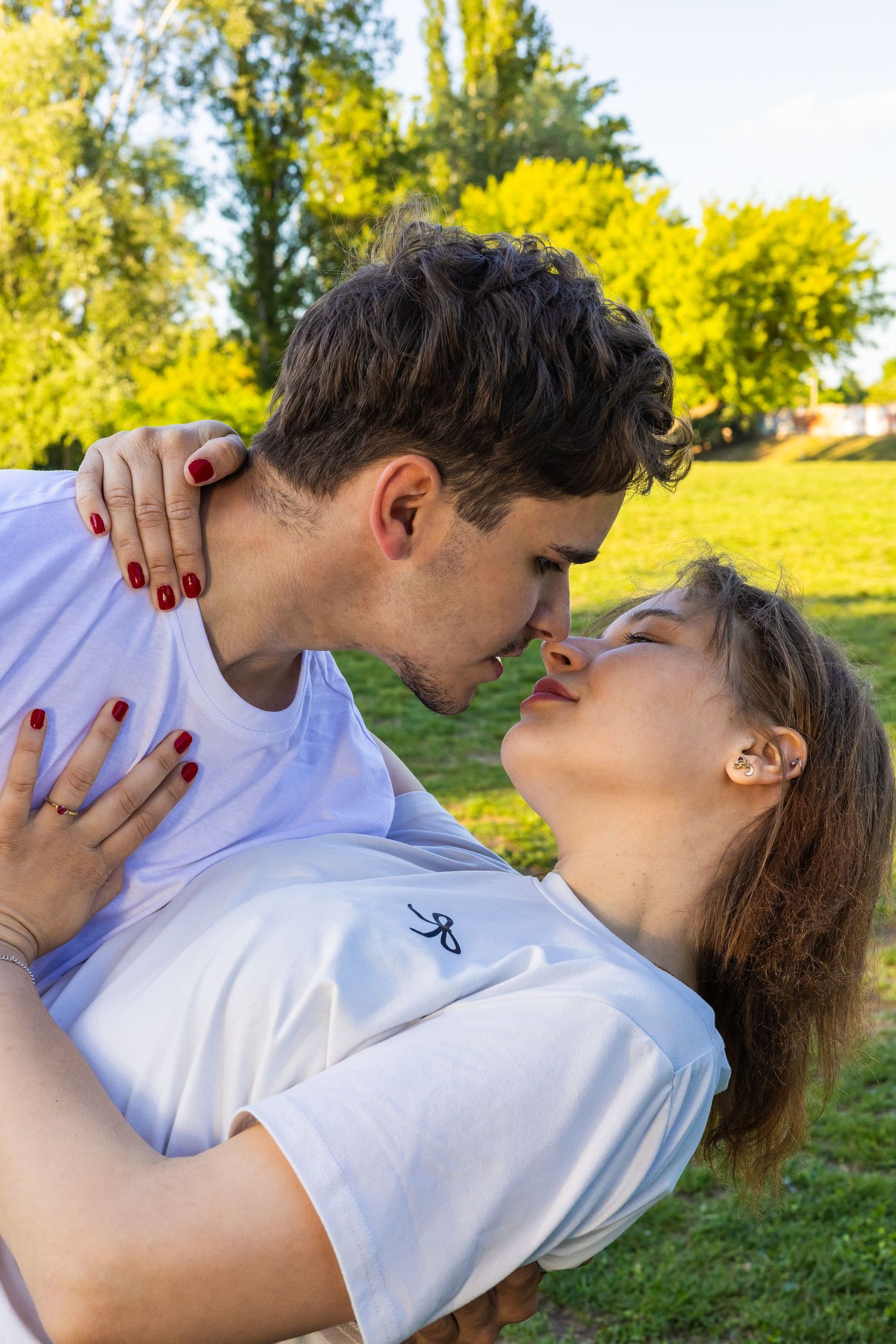 Romantic couple in white t-shirts sharing a kiss outdoors in a sunny green park setting.