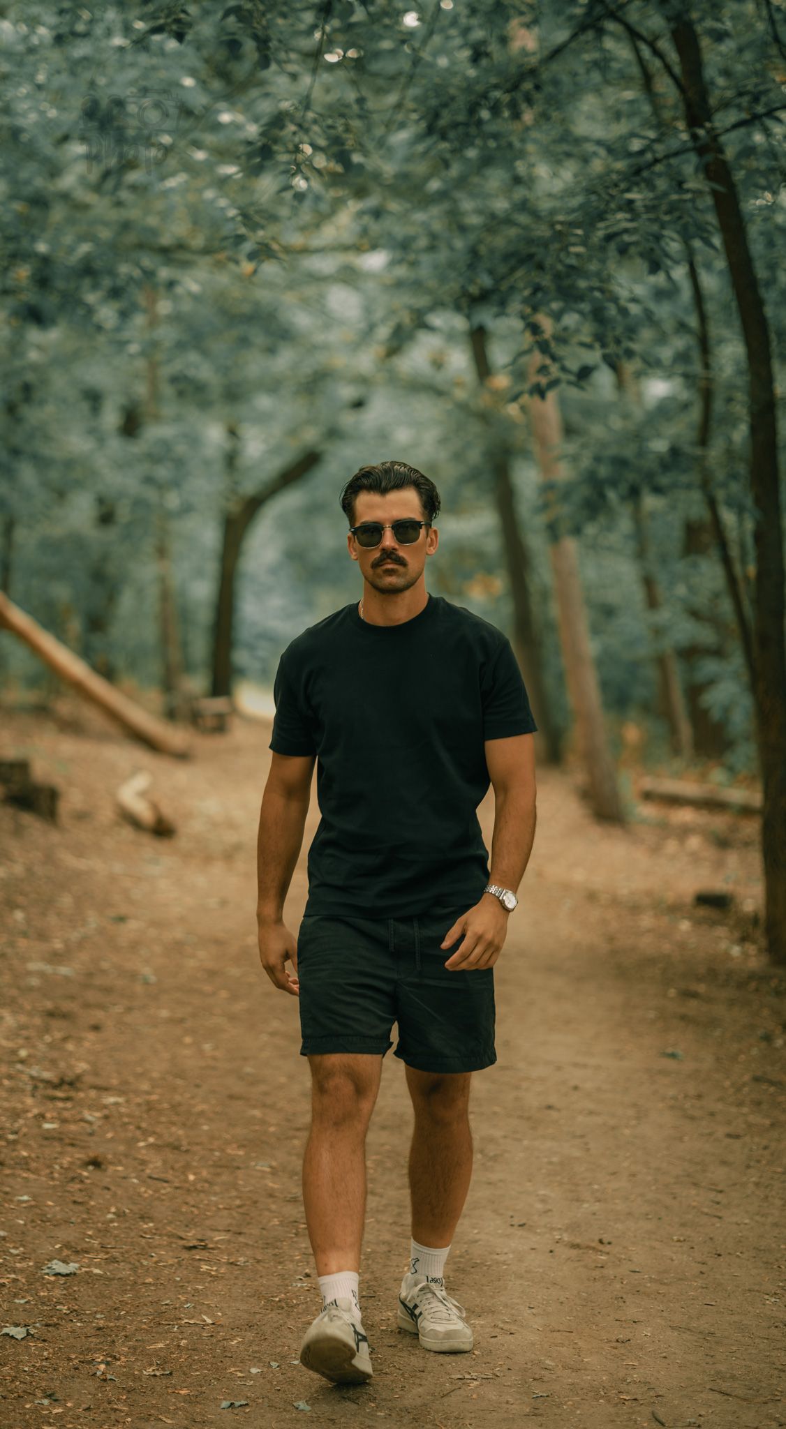 Young man with mustache and sunglasses walking on a forest path wearing black shorts and t-shirt.