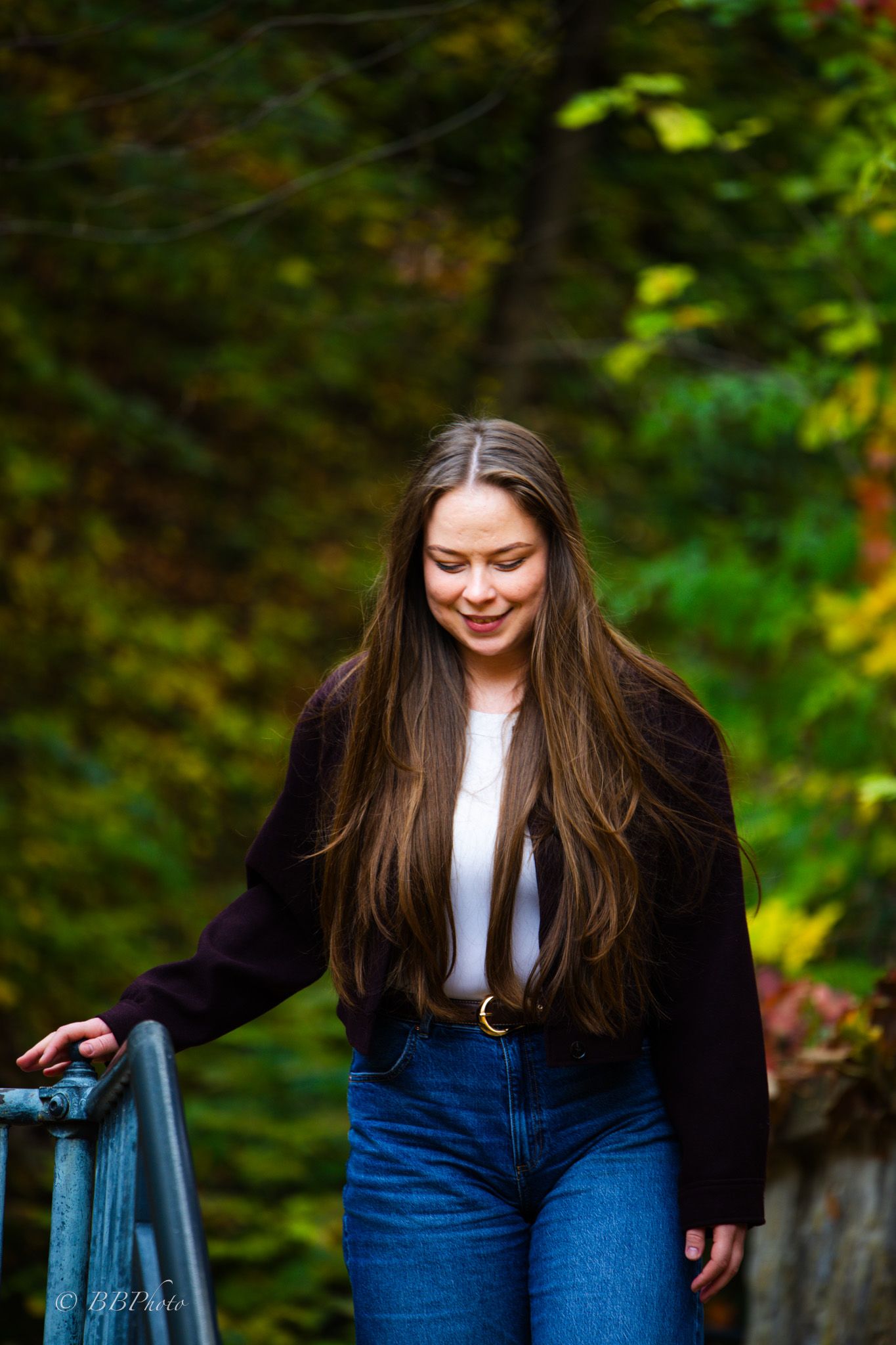 Smiling young woman with long brown hair in a dark cardigan and jeans walks down a railed path surrounded by colorful autumn foliage.