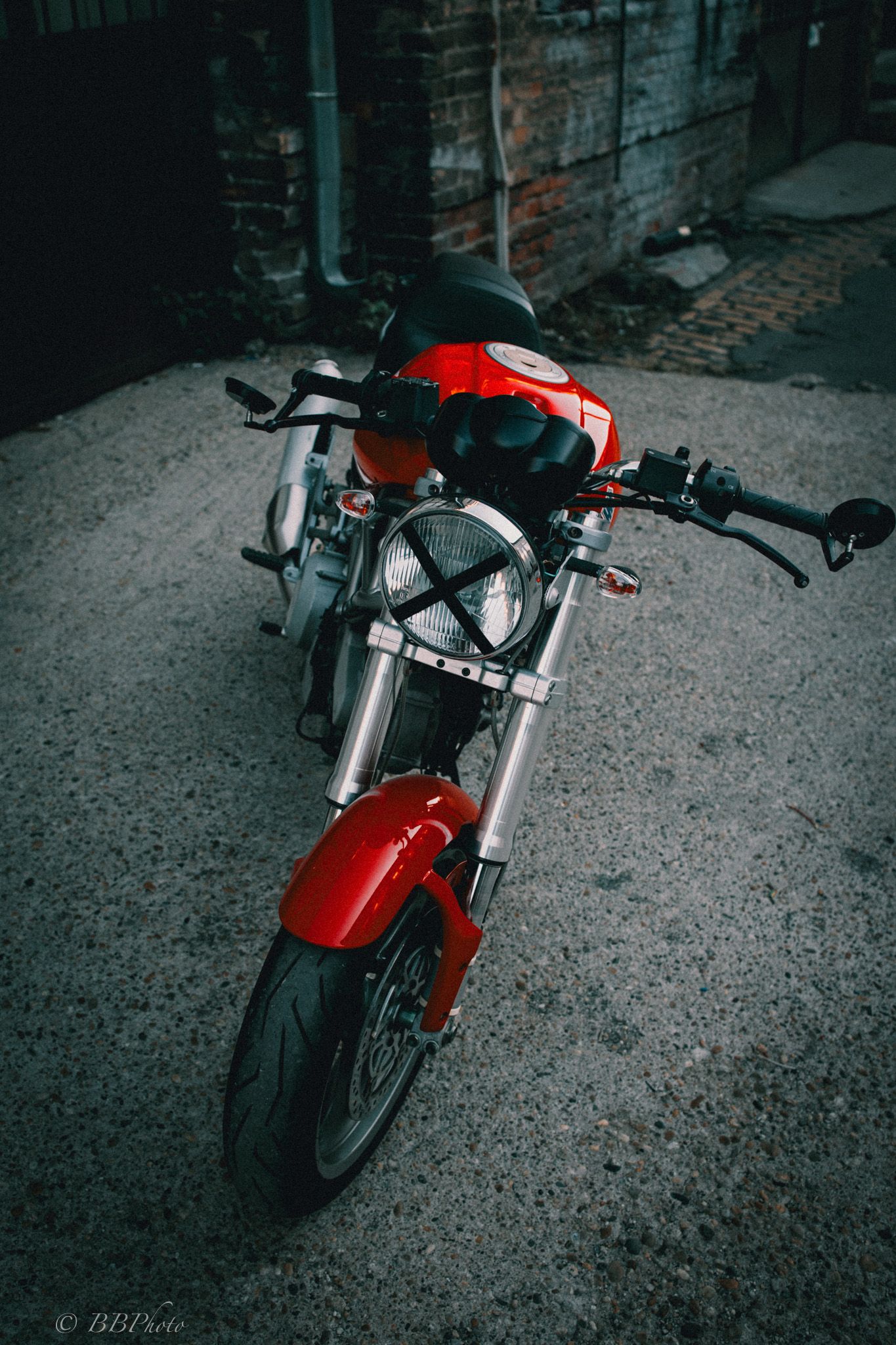 Red naked motorcycle with round headlight and taped X parked on a gray concrete alley beside an old brick wall.