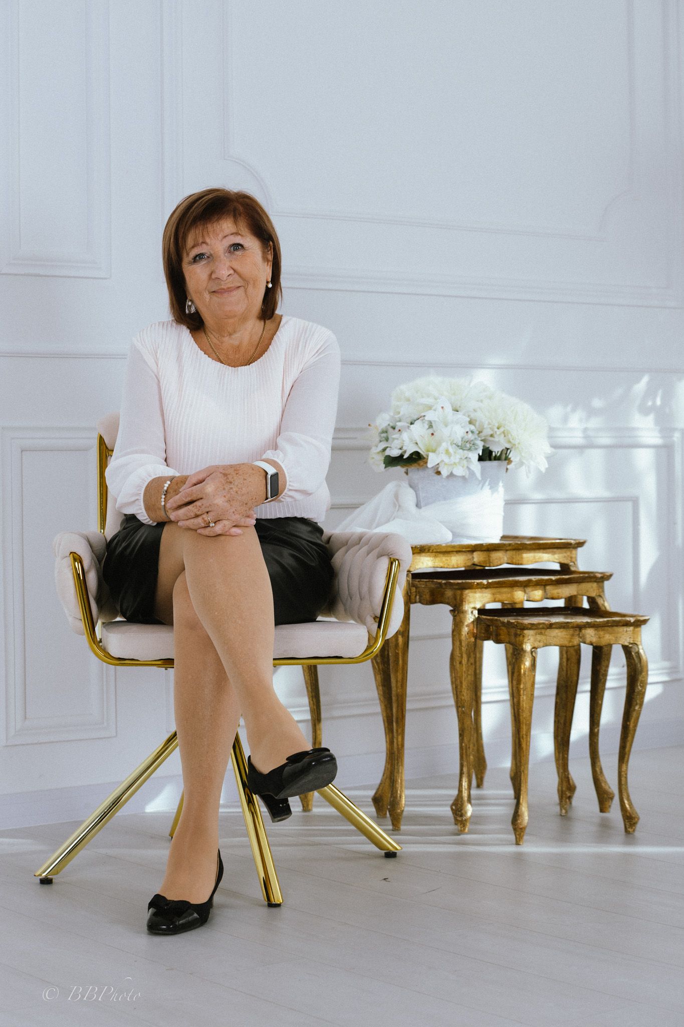 Smiling woman in a white blouse and black skirt sits on a gold-framed upholstered chair beside gilded nesting tables and white flowers.