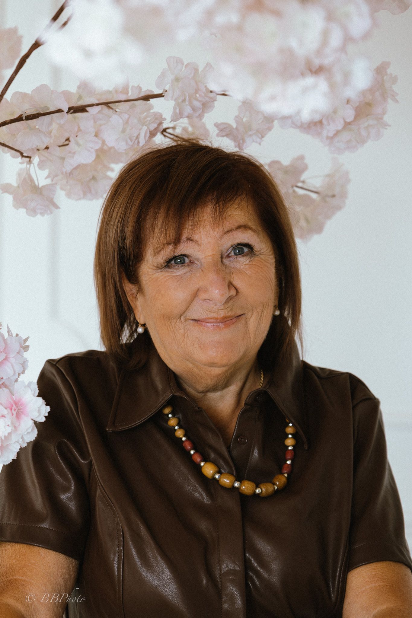 Smiling woman with short brown hair wears a brown leather blouse and amber beaded necklace, seated beside pink cherry blossom branches.
