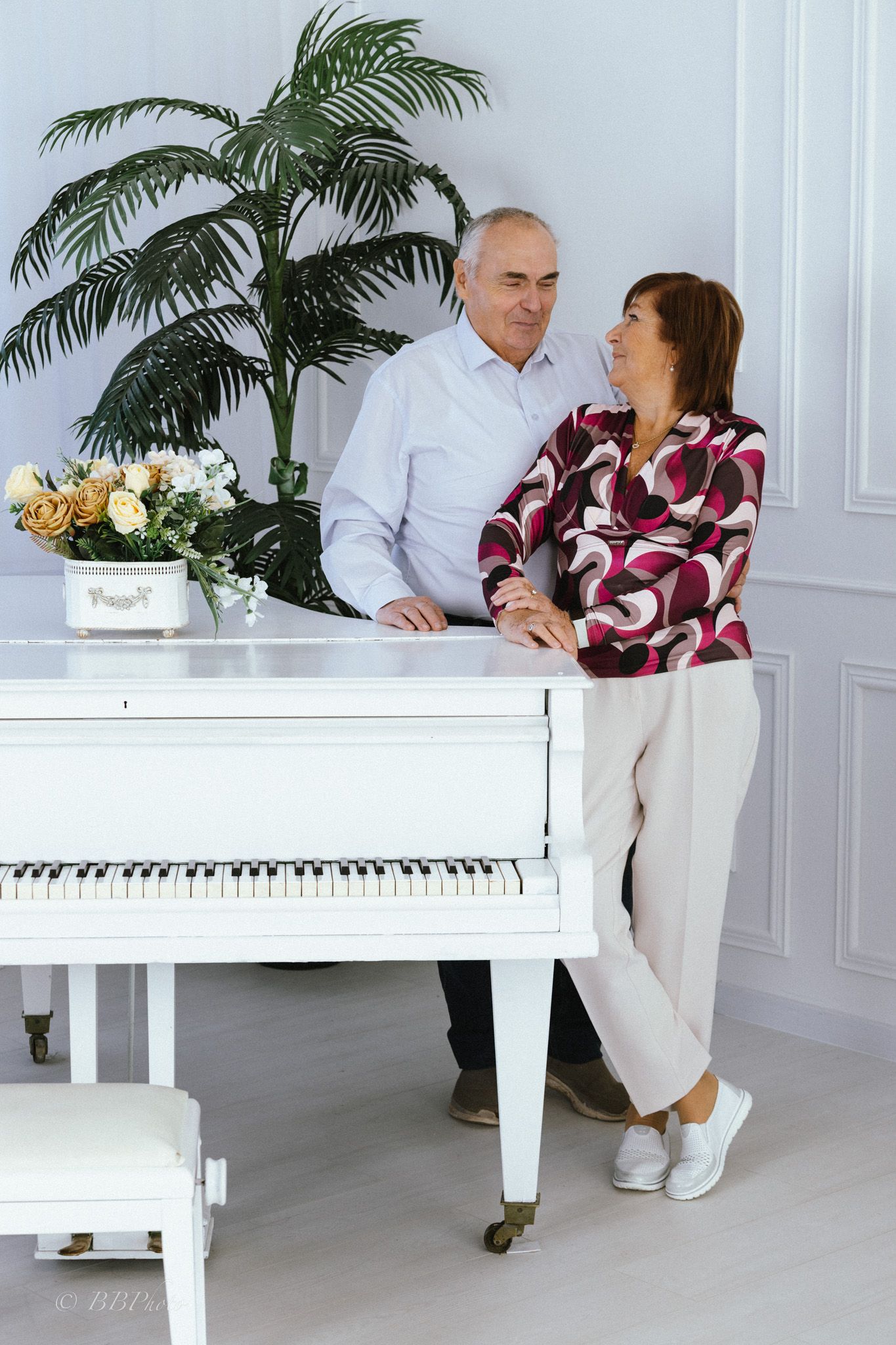 Smiling older couple leaning on a white grand piano beside a potted palm and a bouquet of flowers in a bright, elegant room.