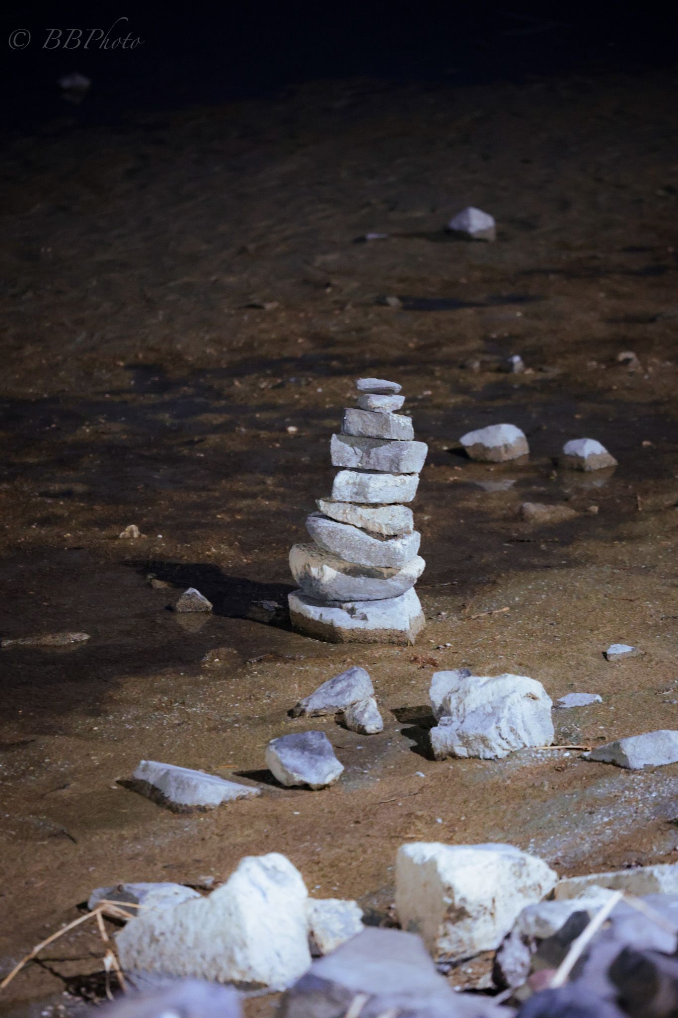 Balanced stack of flat gray stones forming a cairn on sandy ground illuminated in darkness.