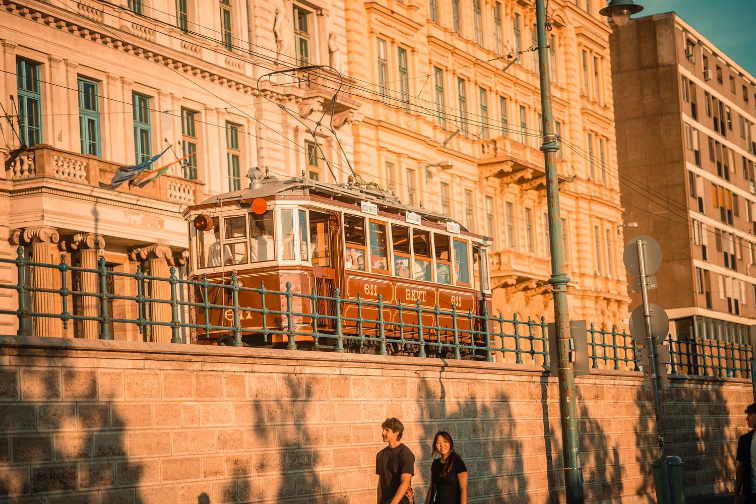 Two people walk beside a historic orange tram passing a grand European building bathed in warm golden sunlight.