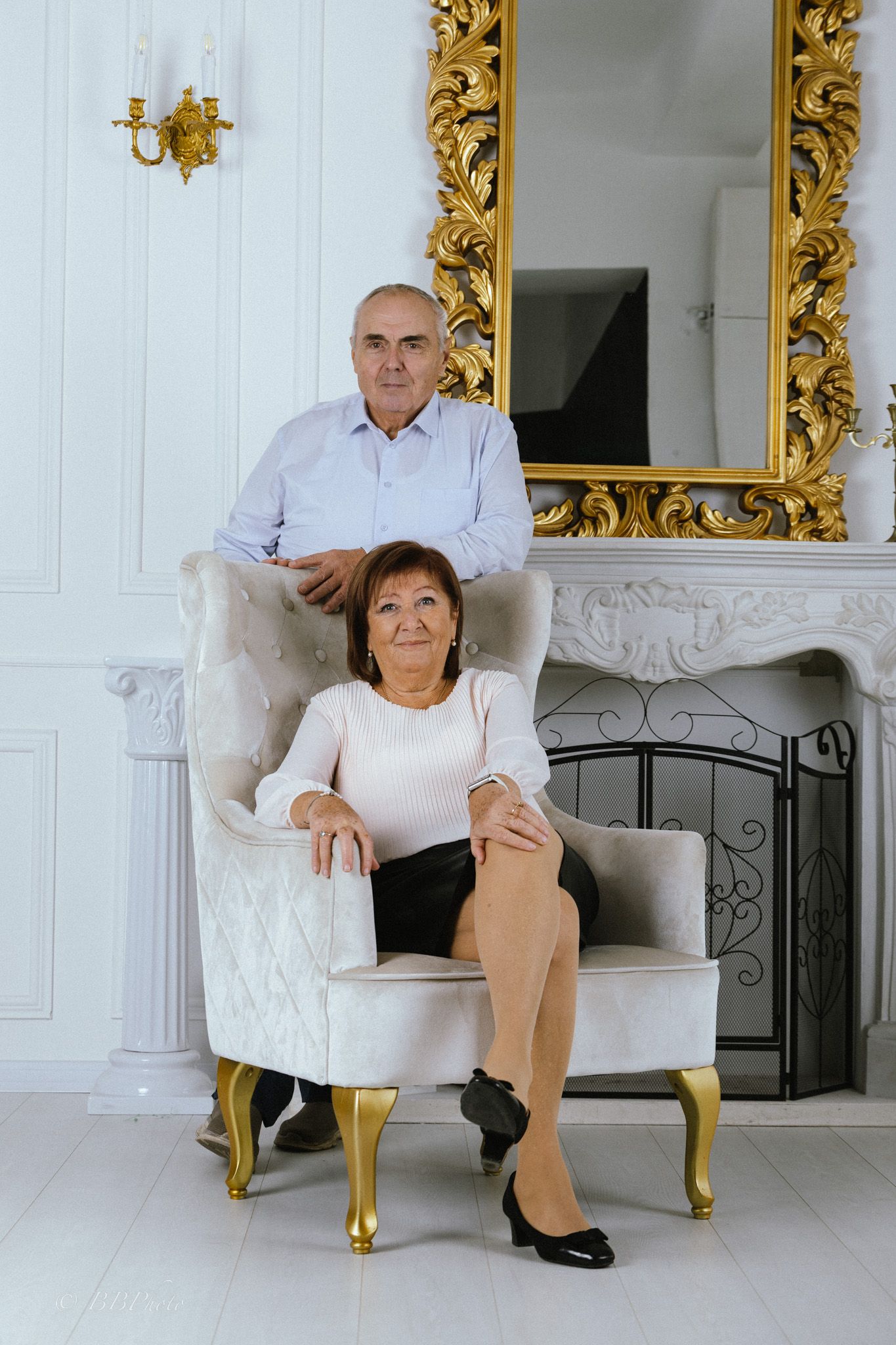 Older couple posing in an elegant room, a woman seated in a tufted armchair and a man standing behind her near a gold-framed mirror.