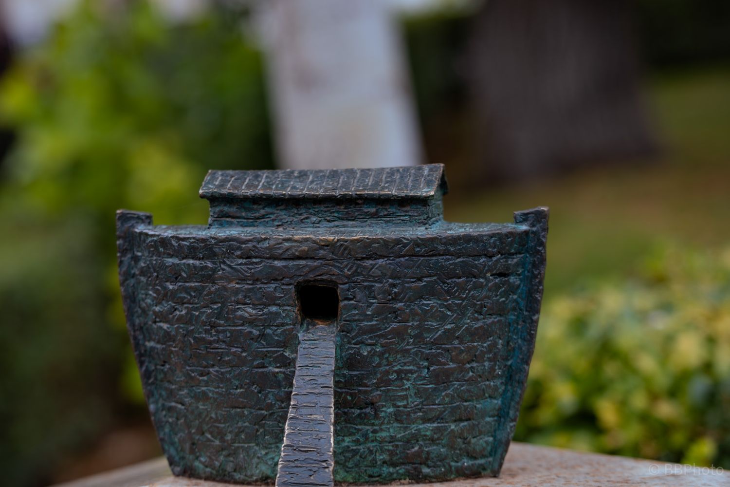 Close-up of an aged bronze miniature boat sculpture with green patina against a blurred garden background.