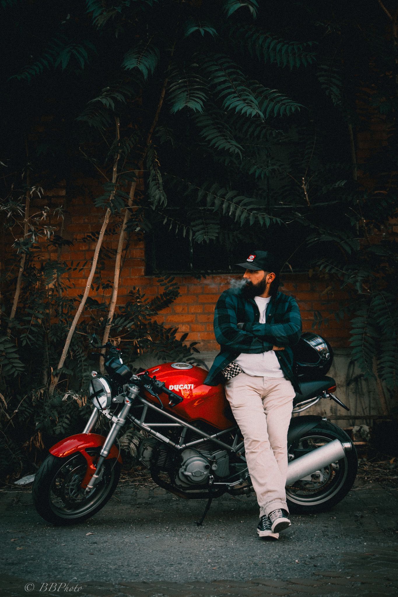 Bearded man in a cap and plaid jacket leans against a red Ducati motorcycle parked in front of a brick wall and green foliage.