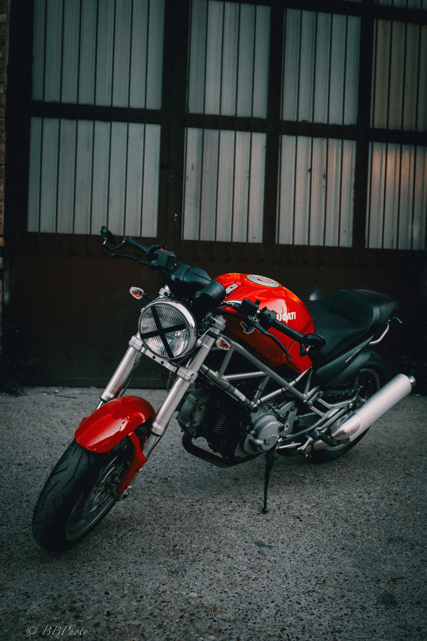 Red Ducati Monster naked motorcycle with exposed trellis frame parked on concrete in front of a dark brown corrugated metal garage door.