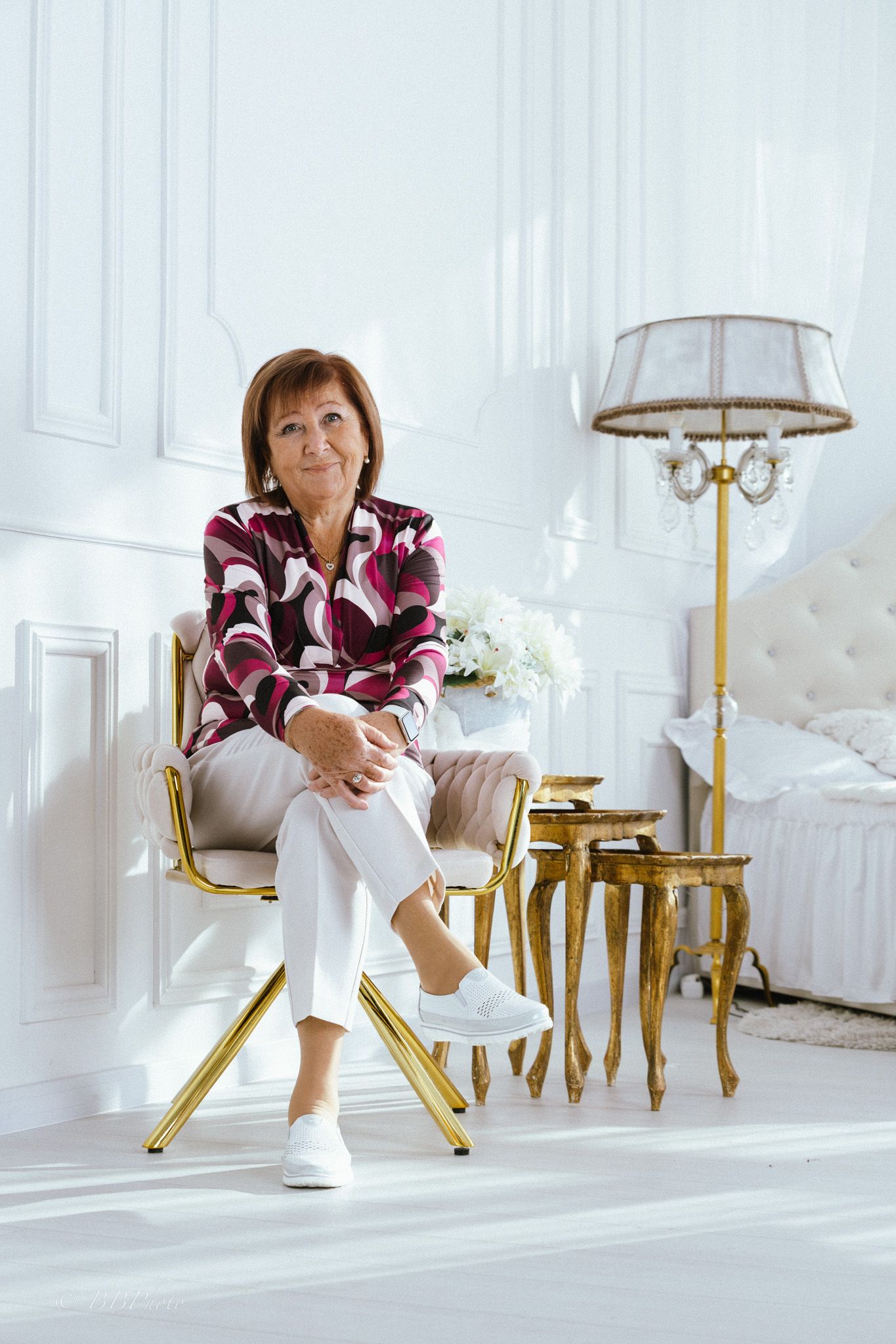 Smiling woman in a patterned jacket and white pants sits on a gold-framed chair in a bright, elegant white bedroom.