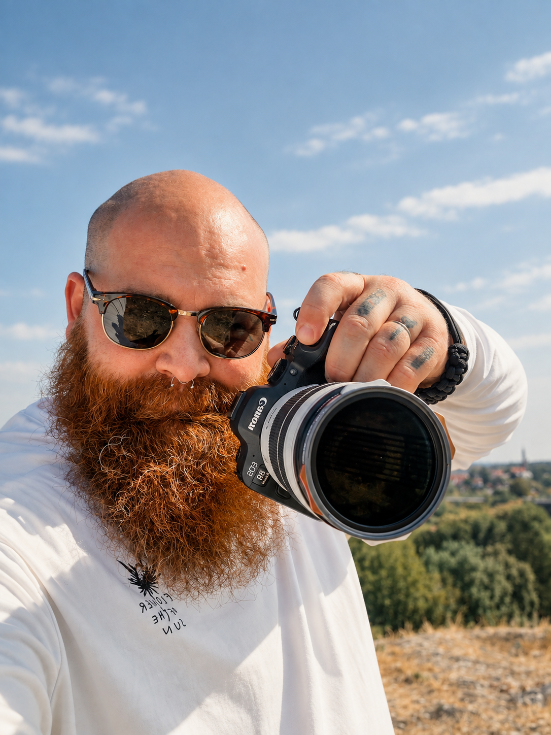Bald man with red beard holding Canon camera outdoors under blue sky, wearing sunglasses and white long-sleeve shirt.