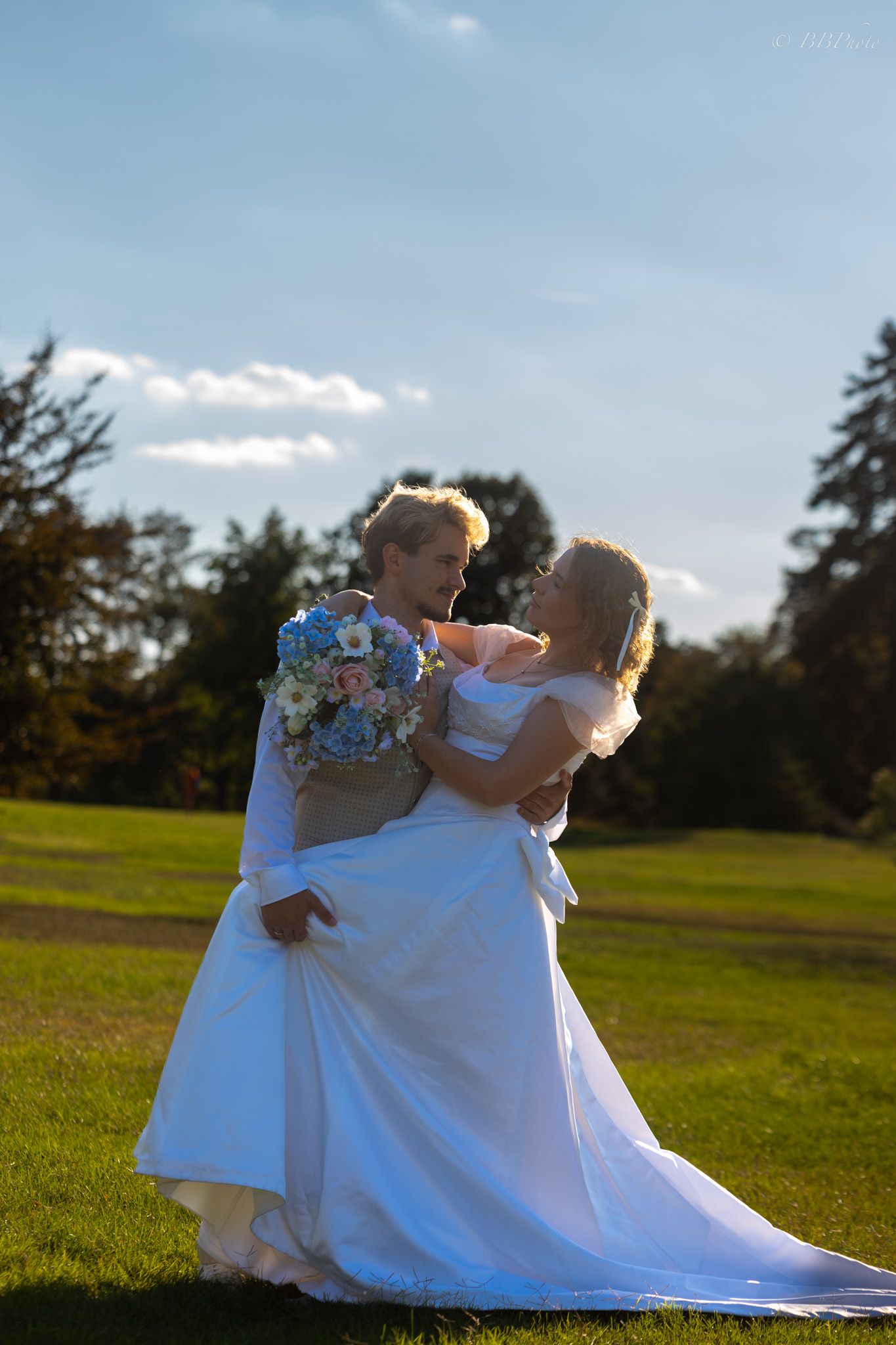 Two brides in white gowns embrace on a sunny green lawn, one holding a blue and white bouquet.