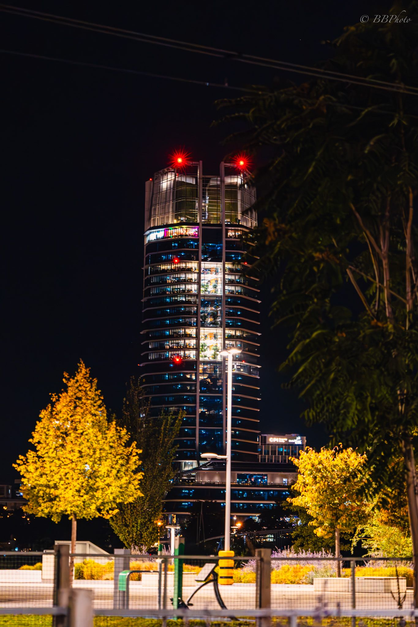Illuminated skyscraper with red lights at night, surrounded by glowing yellow trees and a street lamp in an urban setting.