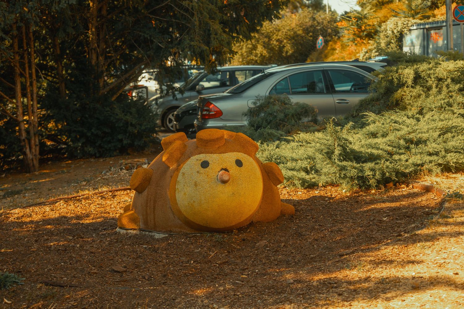 Cute yellow lion character statue lying among fallen autumn leaves in a park near a parking lot.