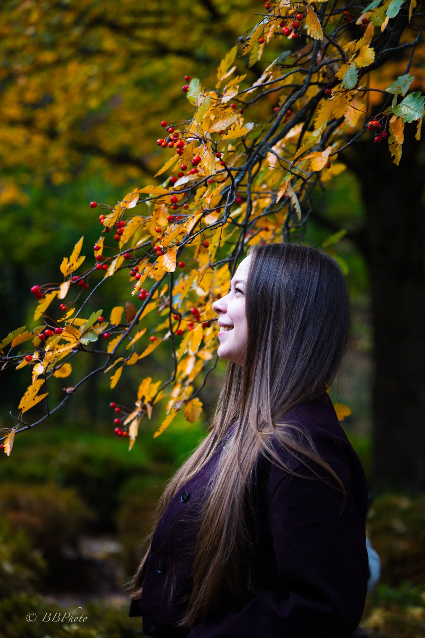 Woman with long brown hair smiles in profile beside autumn branches with golden leaves and red berries in a sunlit park.