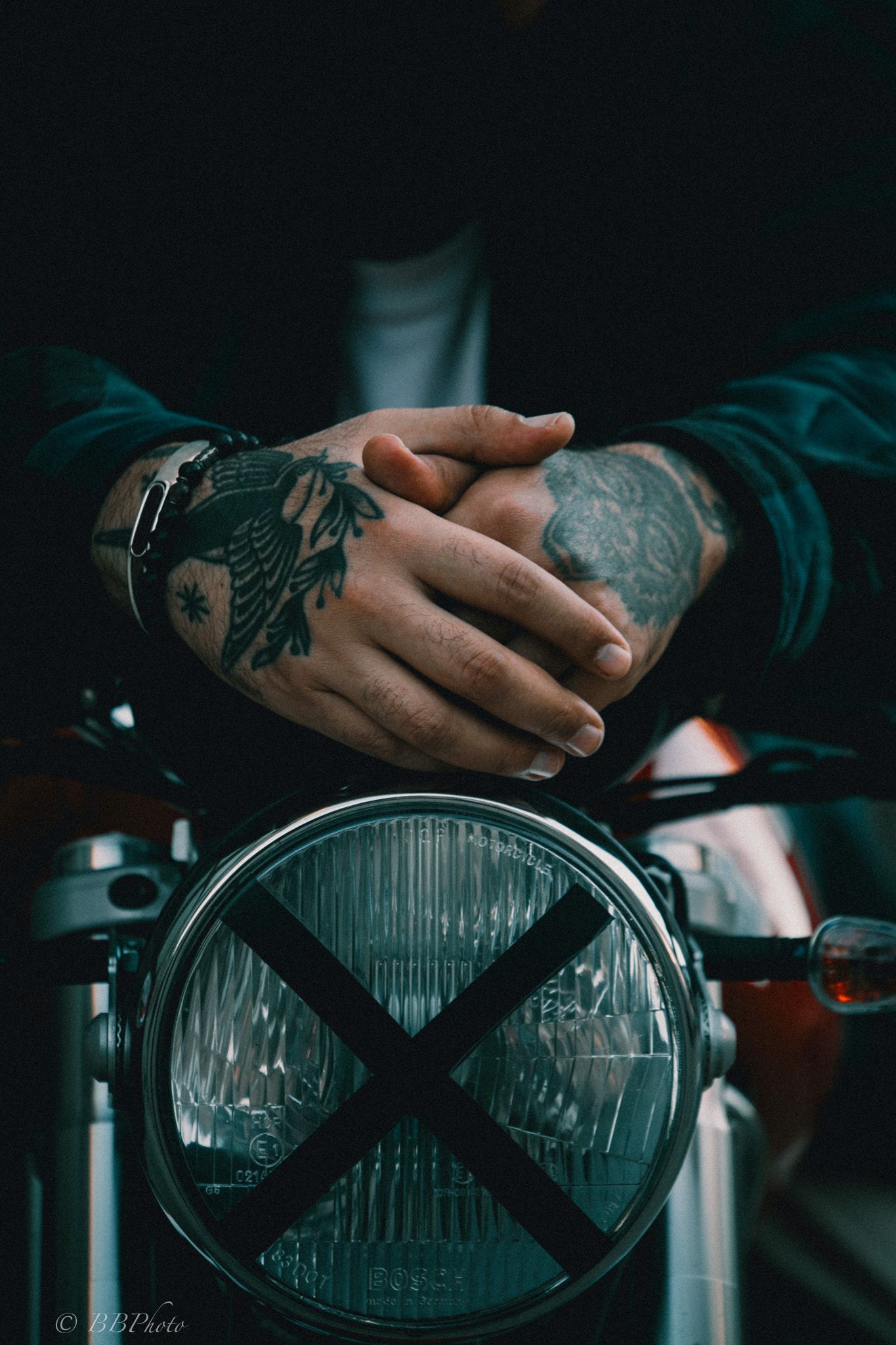 Tattooed hands clasped over a vintage motorcycle headlight marked with a black X, worn by a rider in a dark jacket.