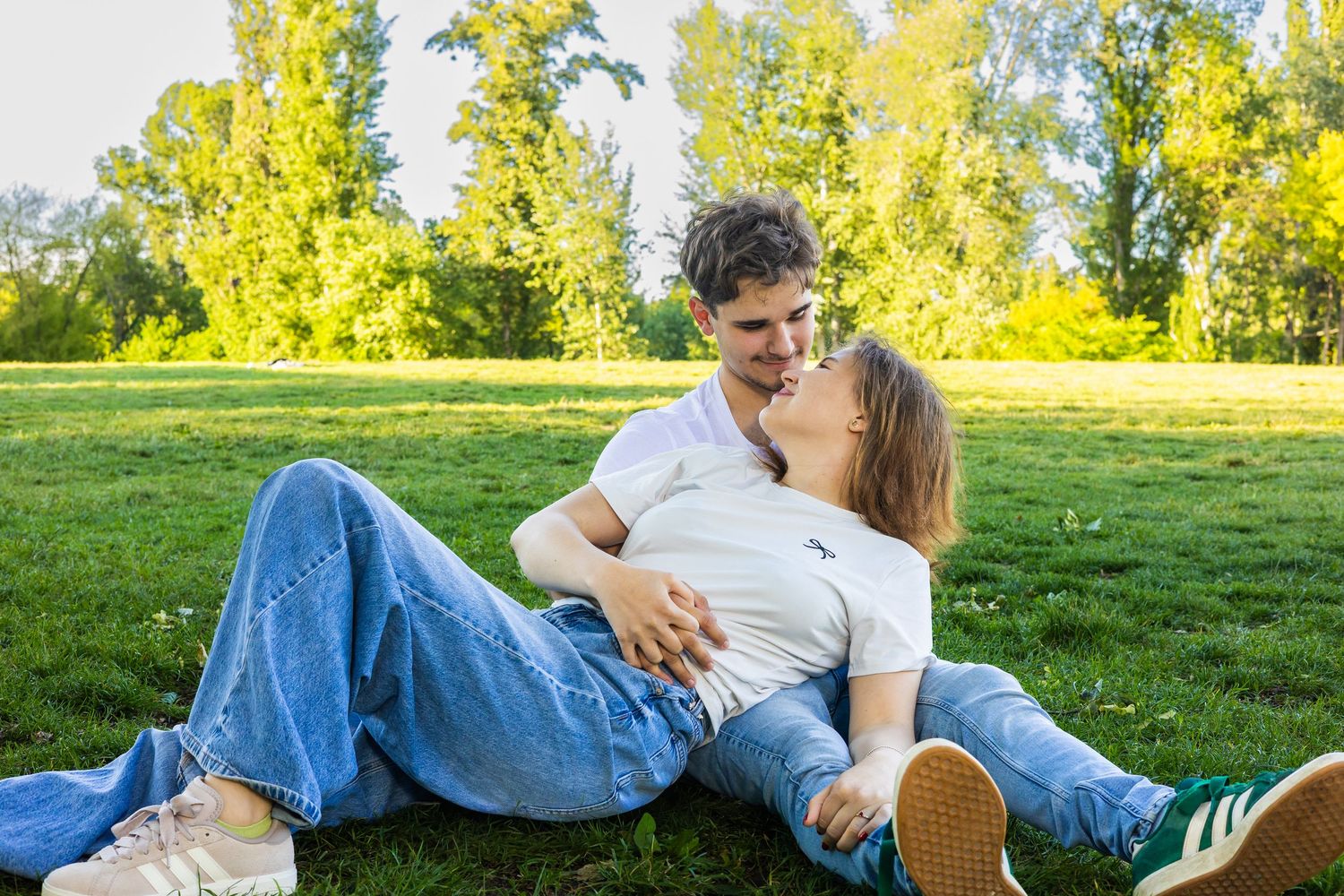 A young couple relaxes on green grass in a sunny park, gazing at each other affectionately.