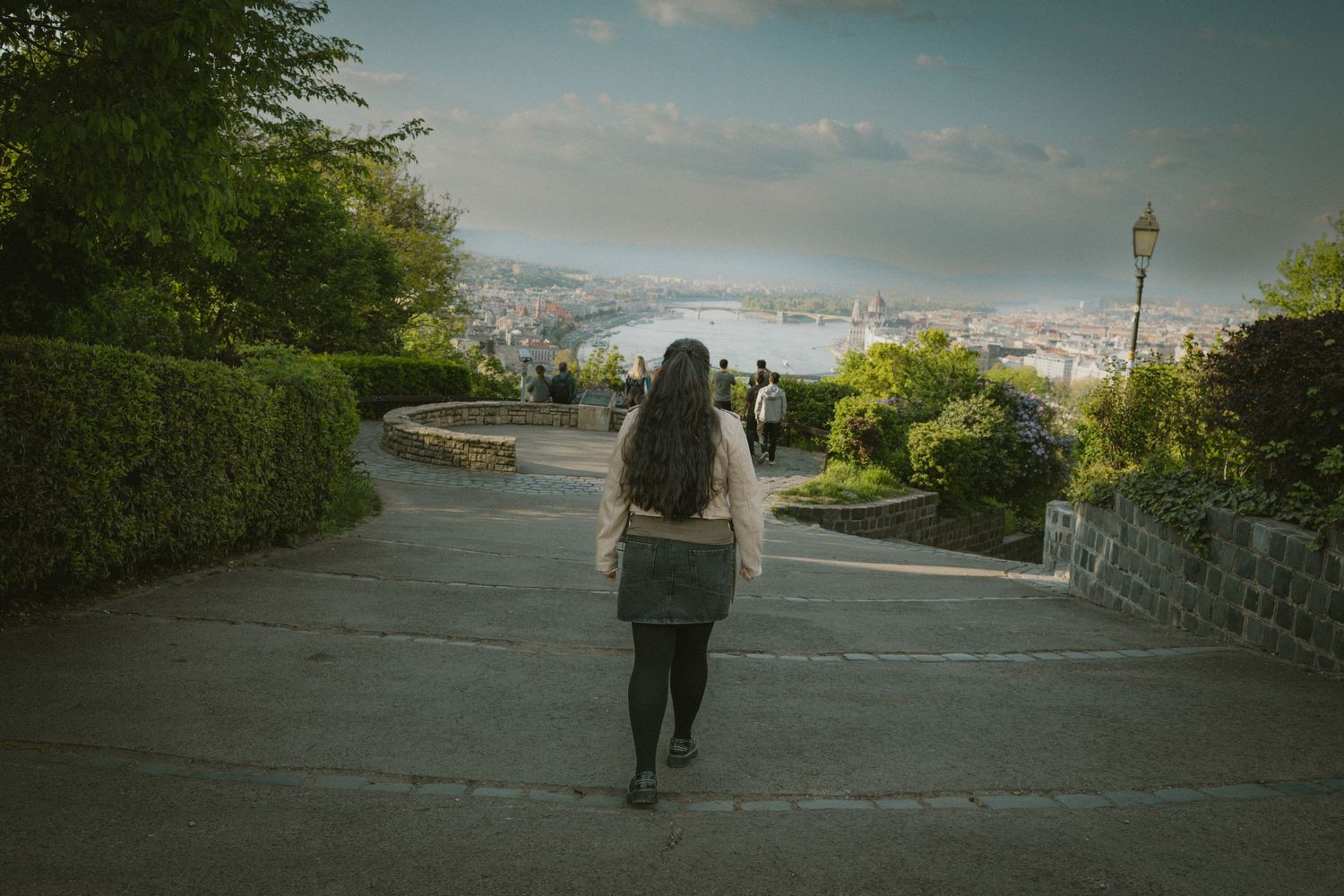 Woman with long dark hair walking along a scenic hilltop park path overlooking a European city panorama.