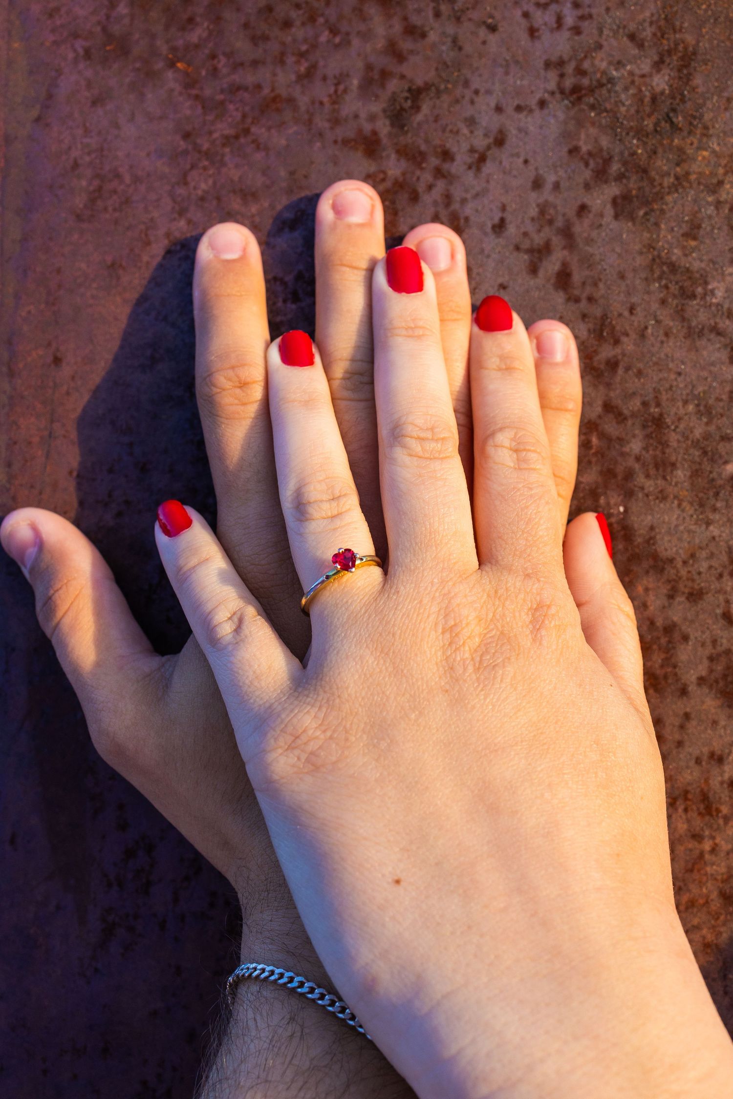 Two hands resting together, one with red nails and a gold ring with red gemstone, on a rustic surface.