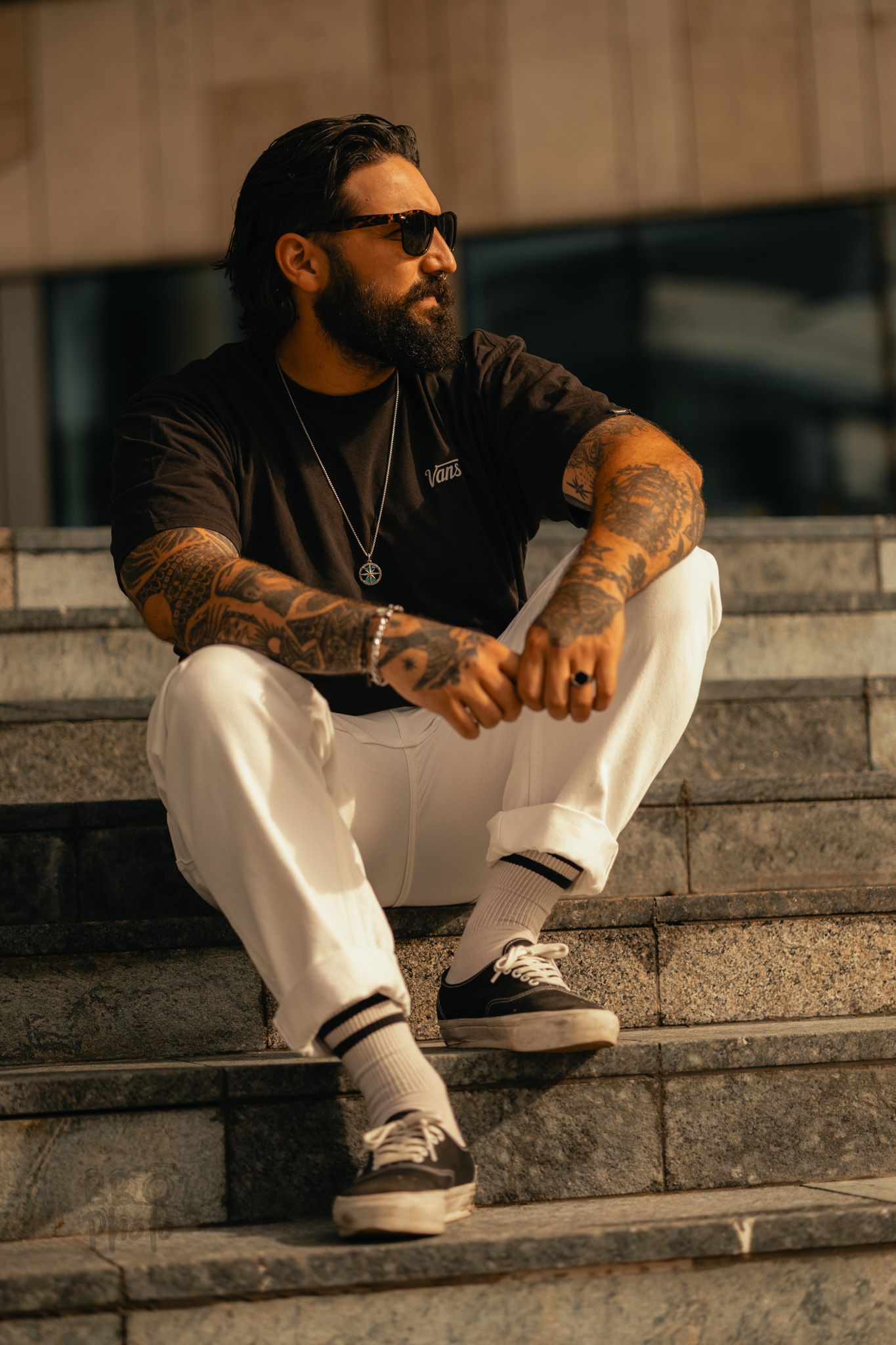 Tattooed man with beard and sunglasses sitting on stone steps wearing black tee, white pants, and dark sneakers.