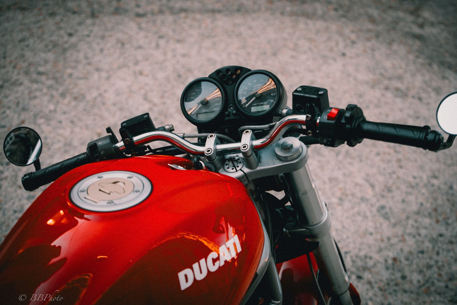 Close-up of a red Ducati motorcycle fuel tank, handlebars, and analog gauges, viewed from the rider's seat on a gravel surface.