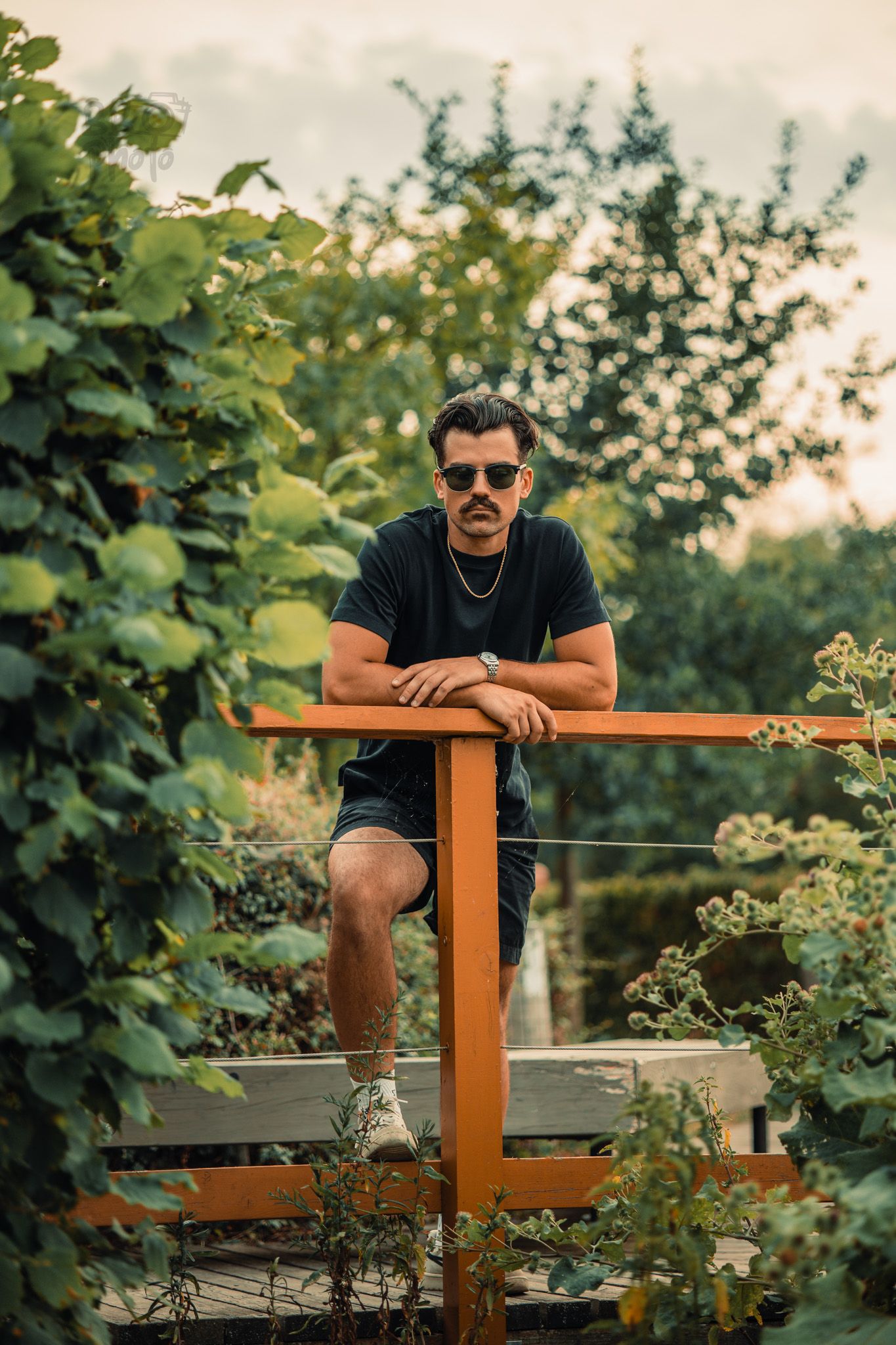 Man with sunglasses and mustache leaning on orange railing in lush green garden at sunset.