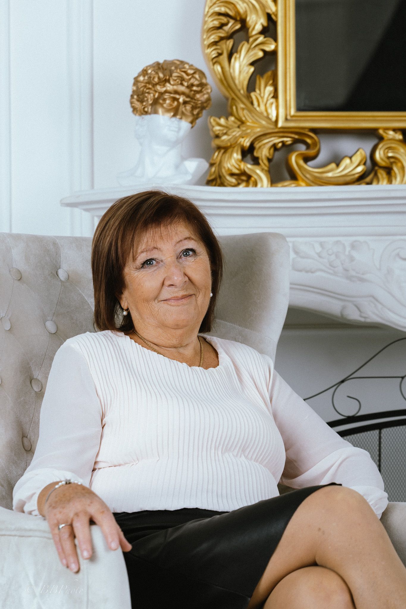 Smiling older woman with short brown hair sits in a tufted gray armchair beside an ornate white fireplace with a gilded mirror.