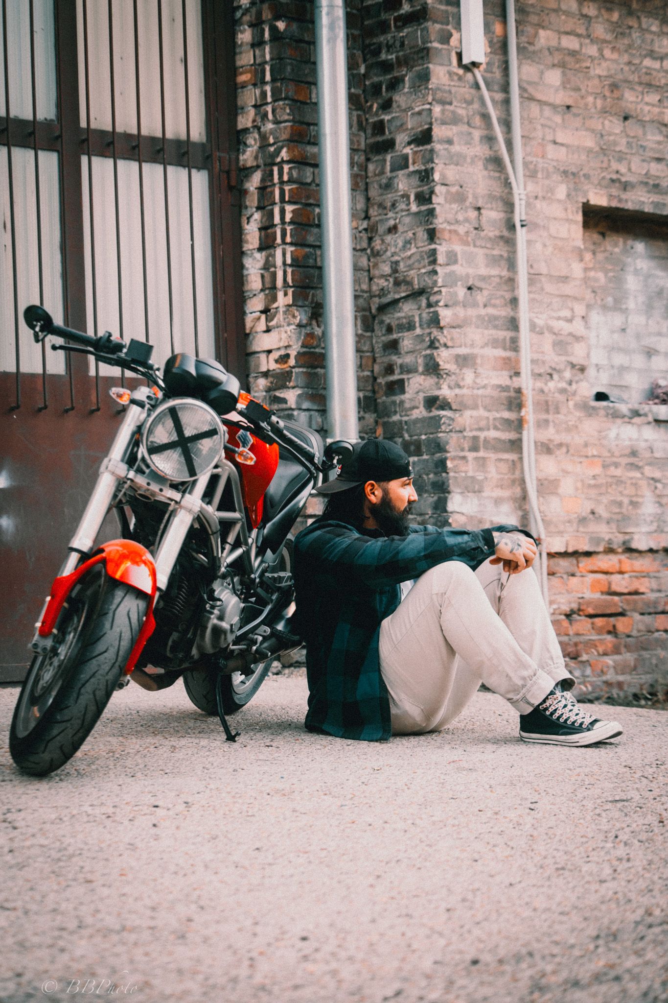 Bearded man in a black cap and plaid shirt sits on the ground beside a red and black motorcycle in front of a rustic brick wall.