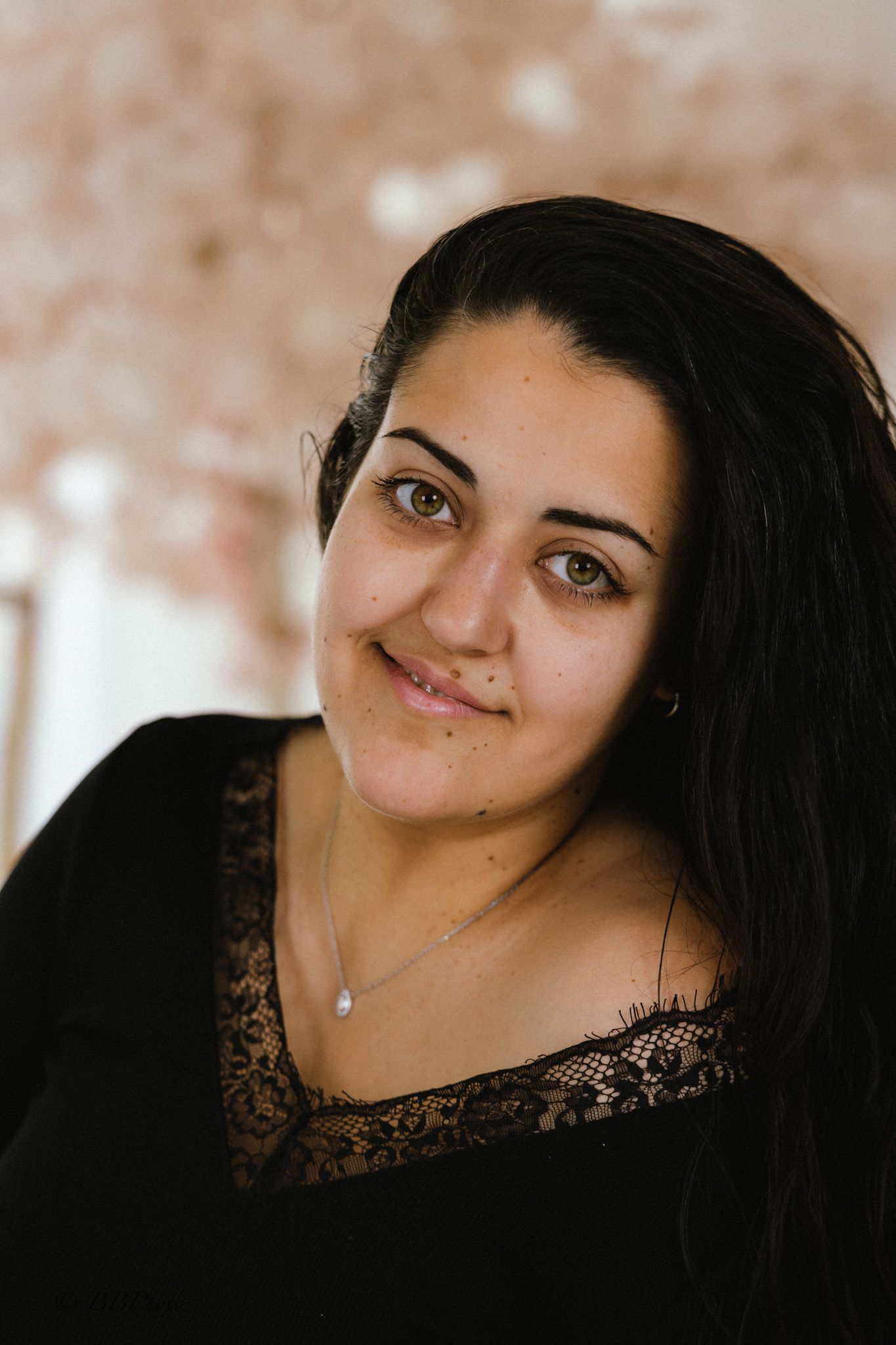 Smiling woman with long dark hair wearing a black lace-trim top and a delicate pendant necklace in a bright, soft-toned indoor portrait.