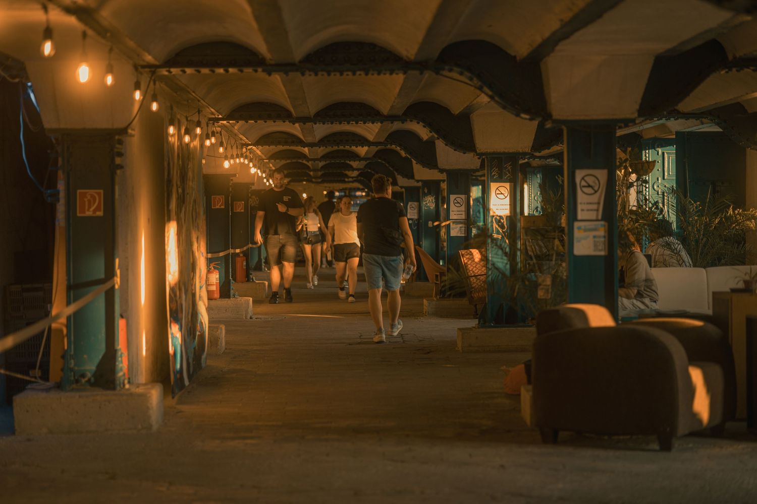 Dimly lit covered walkway with string lights, shops on both sides, and people strolling through a warm evening marketplace.