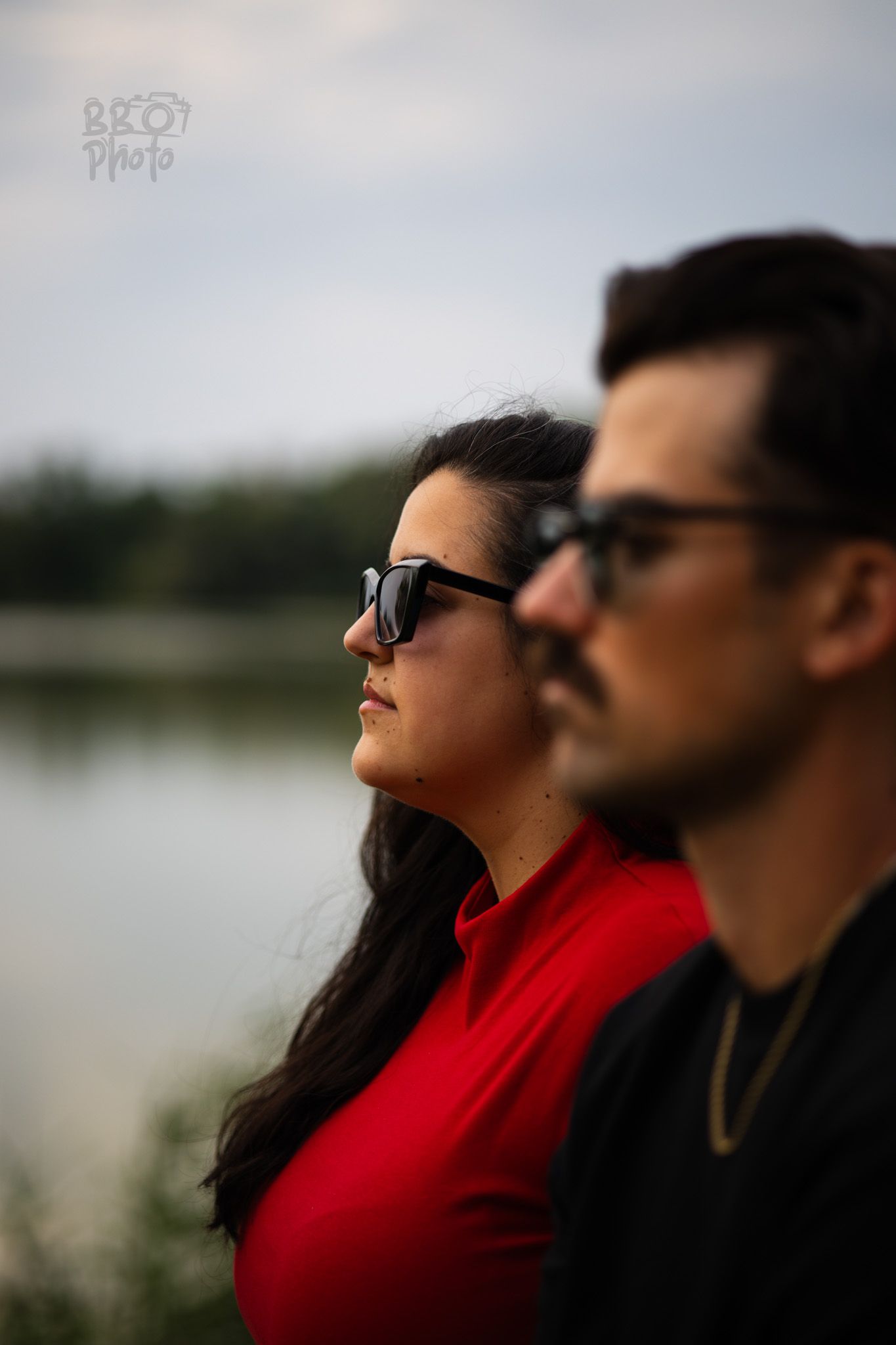 Woman in red top and man in black shirt both wearing sunglasses gazing into the distance by a lake.