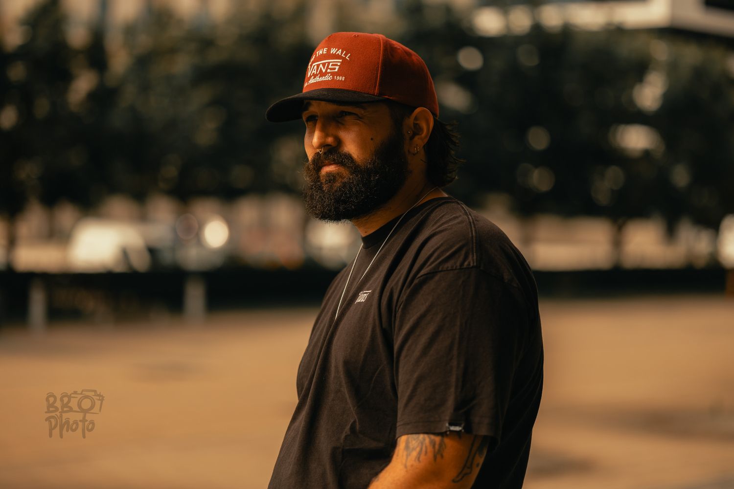 Bearded man in black t-shirt and red trucker hat poses outdoors in urban setting at dusk.