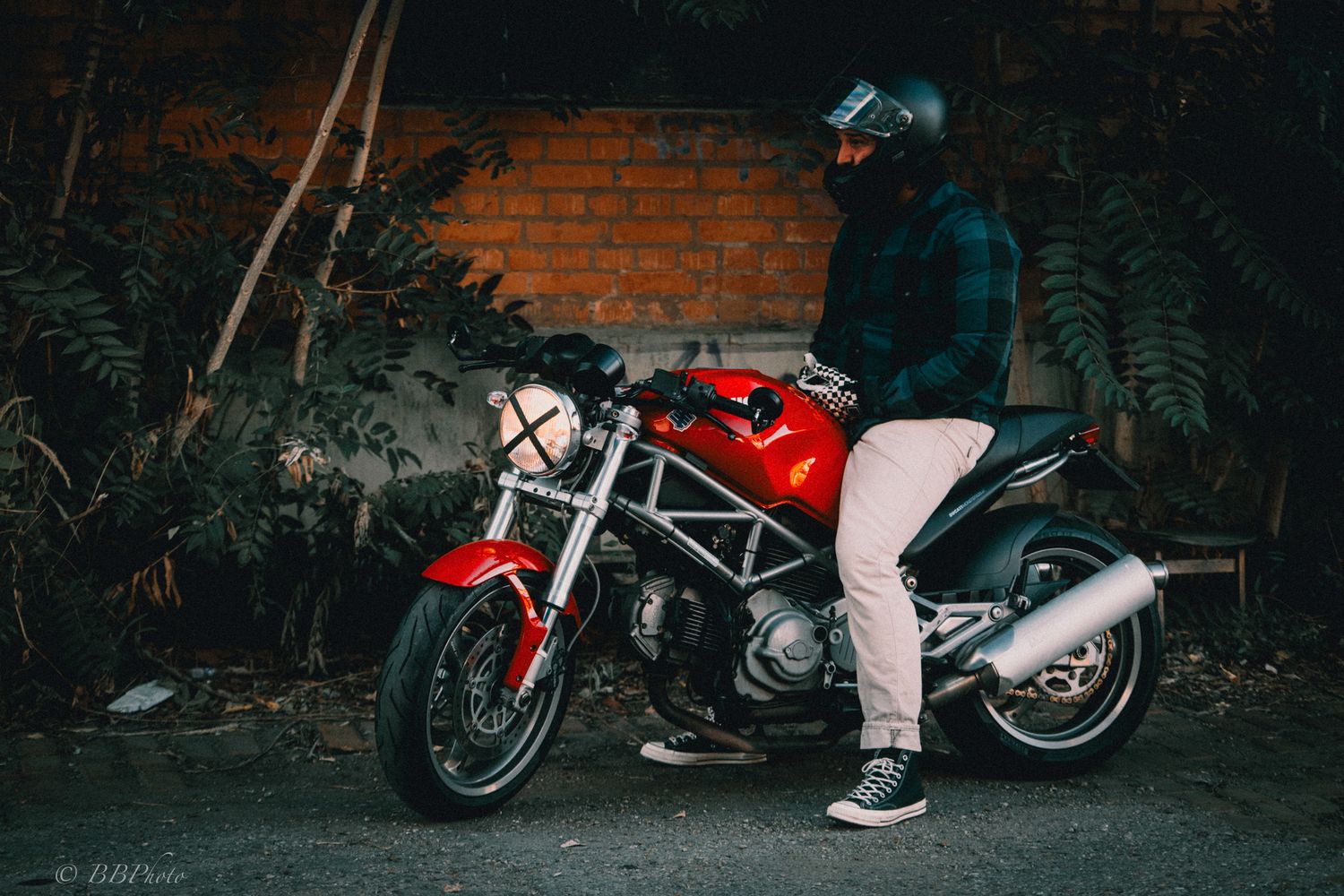 Rider in a helmet and plaid jacket sits on a red naked sport motorcycle parked in front of a brick wall with green fern plants.