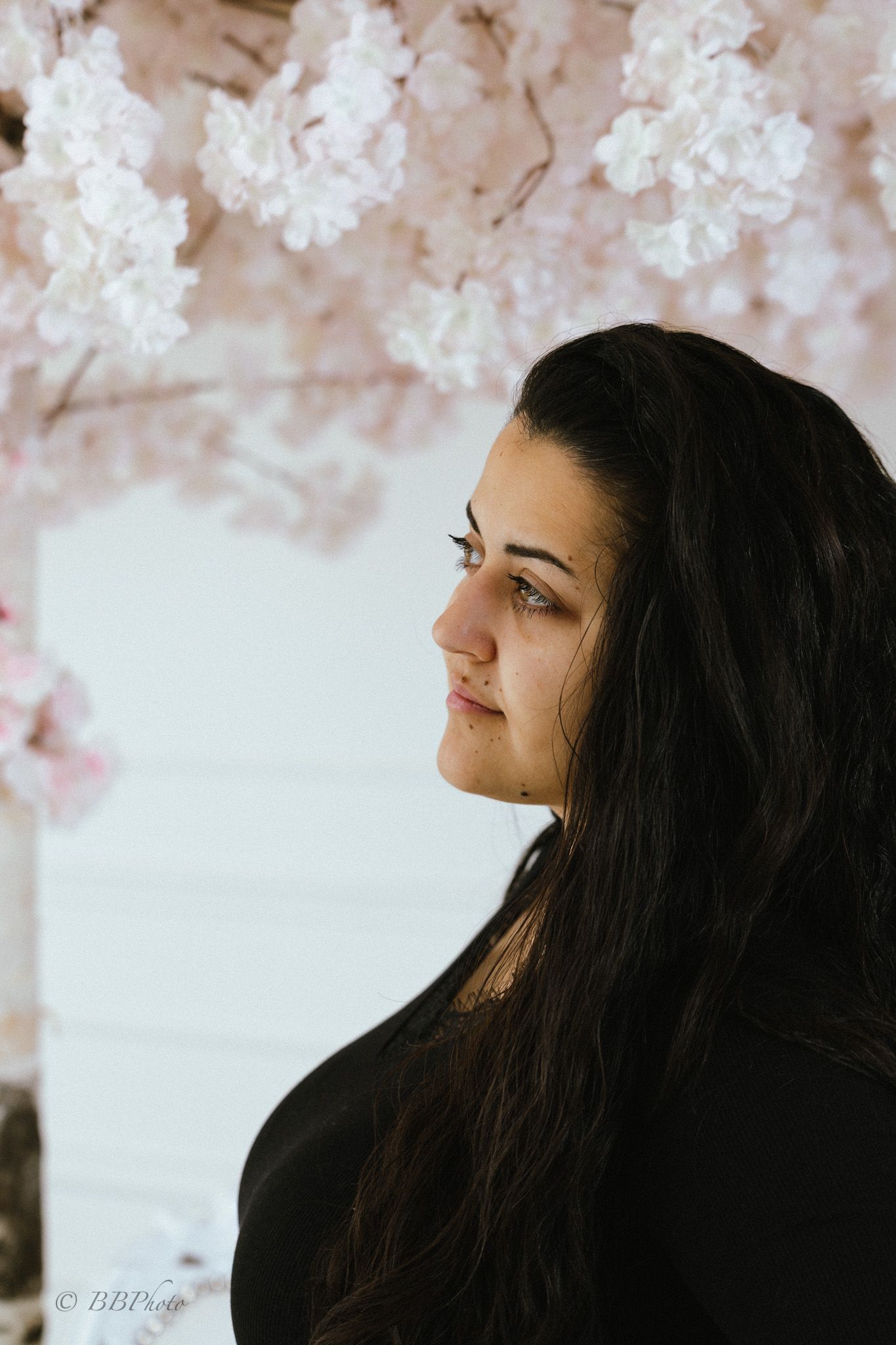 Side profile portrait of a woman with long dark hair in a black top, gazing away beside a wall of soft pink cherry blossom branches.