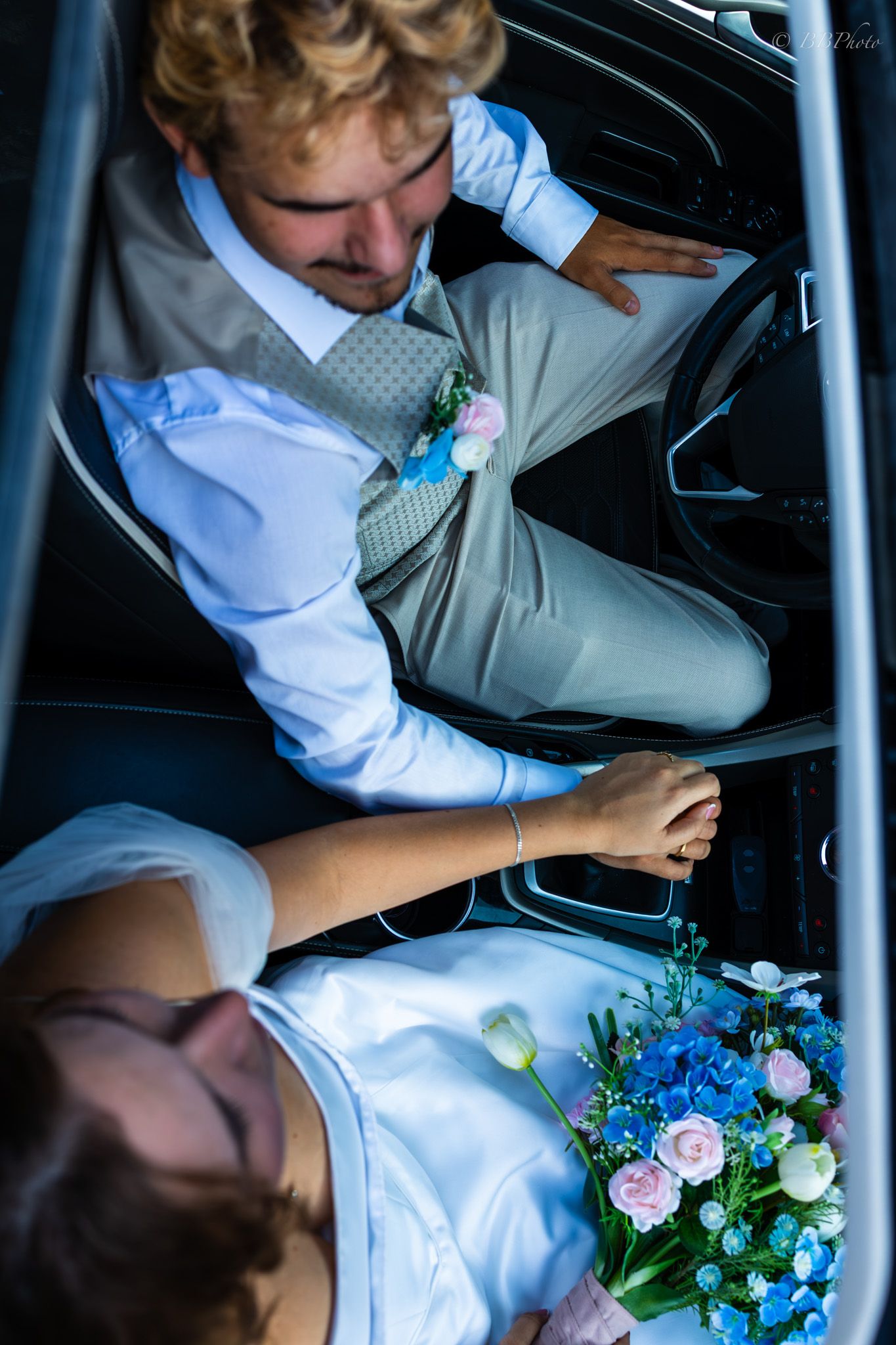Aerial view of a bride and groom sitting in a car, bride holding blue and pink floral bouquet, groom wearing boutonniere.