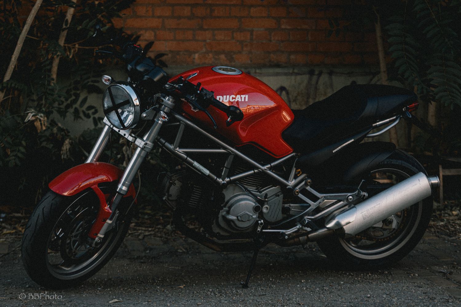 Red Ducati Monster motorcycle with exposed steel trellis frame and chrome exhaust parked on a street beside a brick wall.