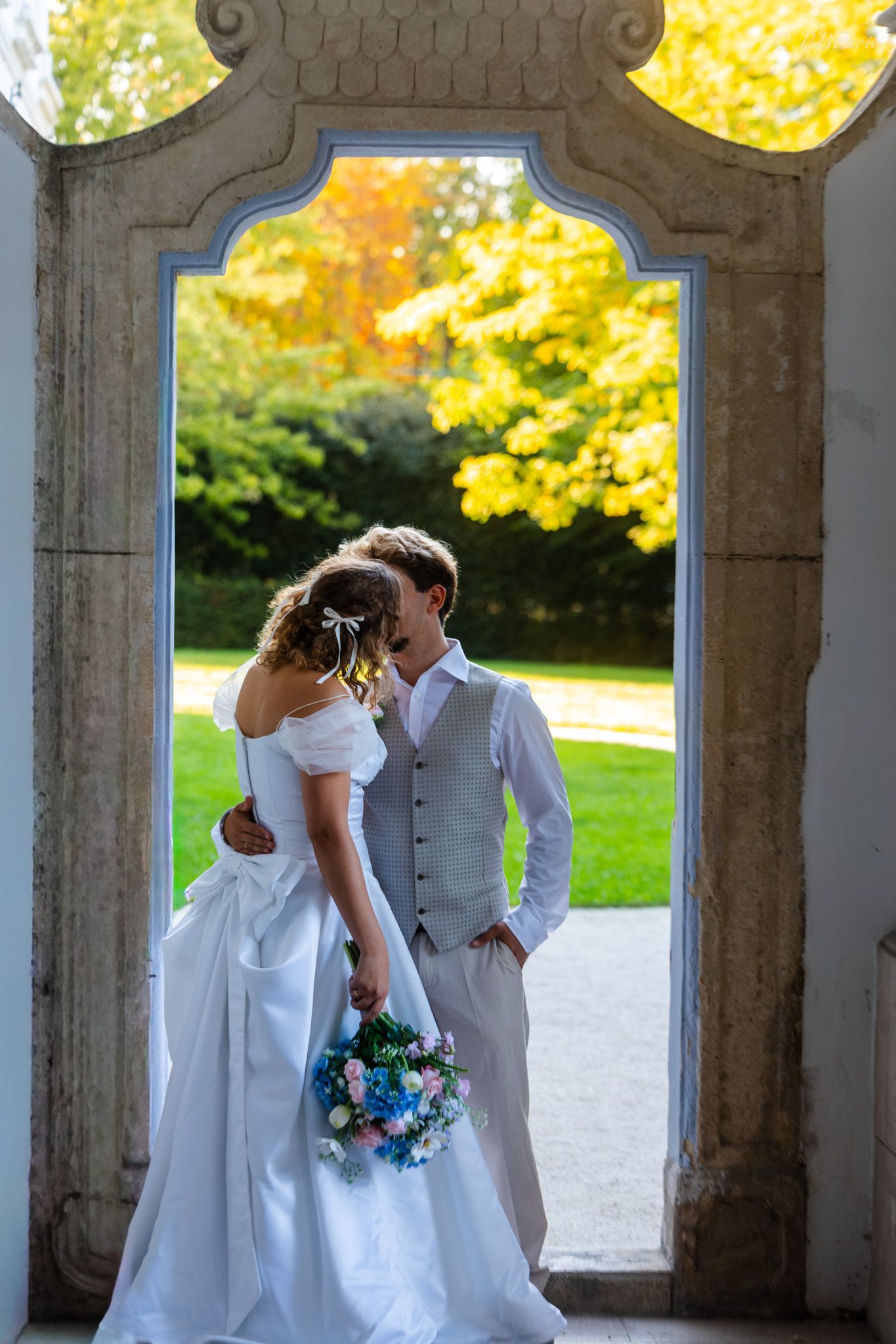 Bride and groom sharing a romantic moment in an ornate archway, surrounded by lush autumn greenery.